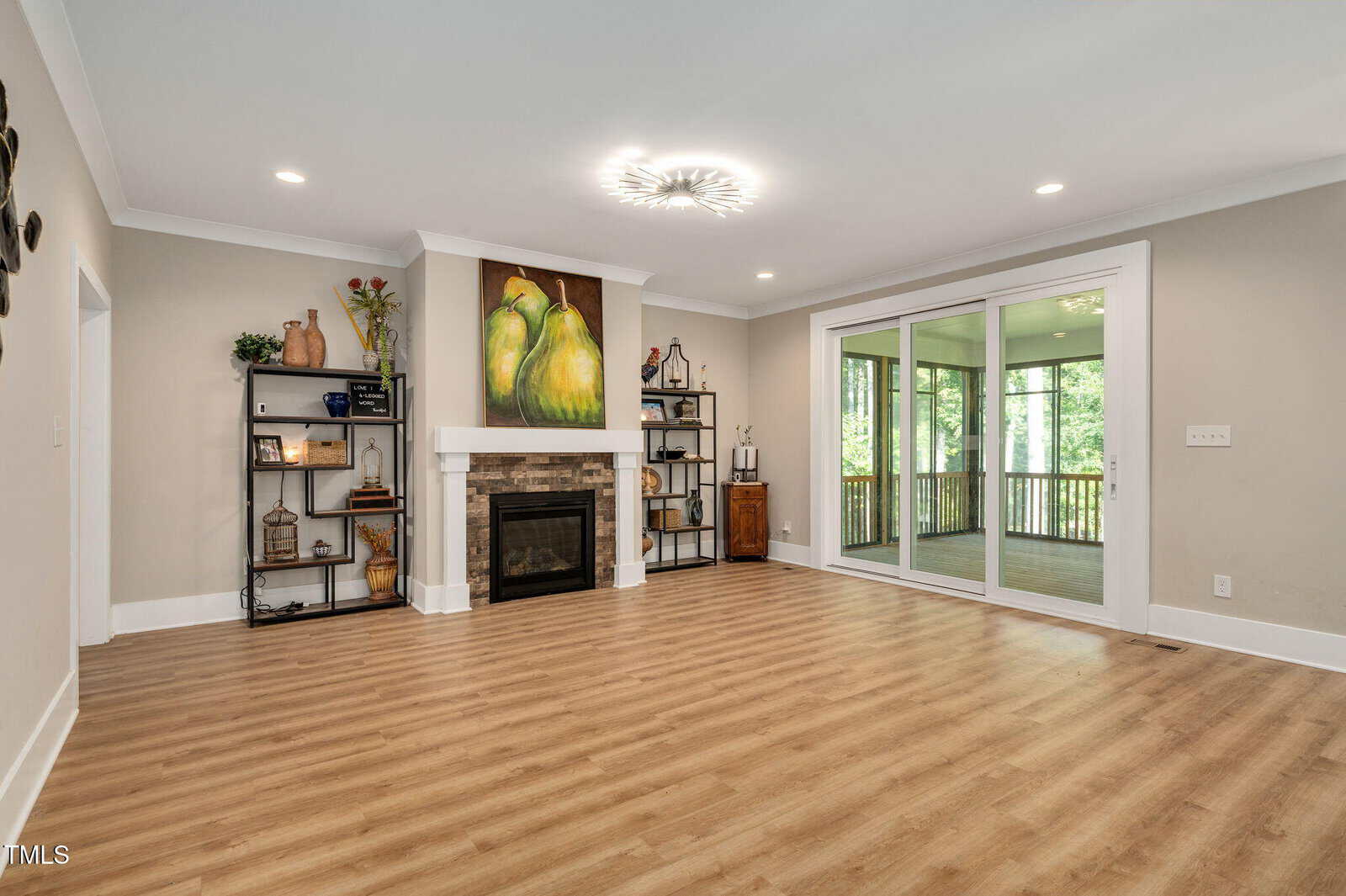 A spacious living room with light wood flooring, a white fireplace with stone accents, shelves on each side, a large painting above the mantel, and sliding glass doors leading to a screened porch with trees visible outside.