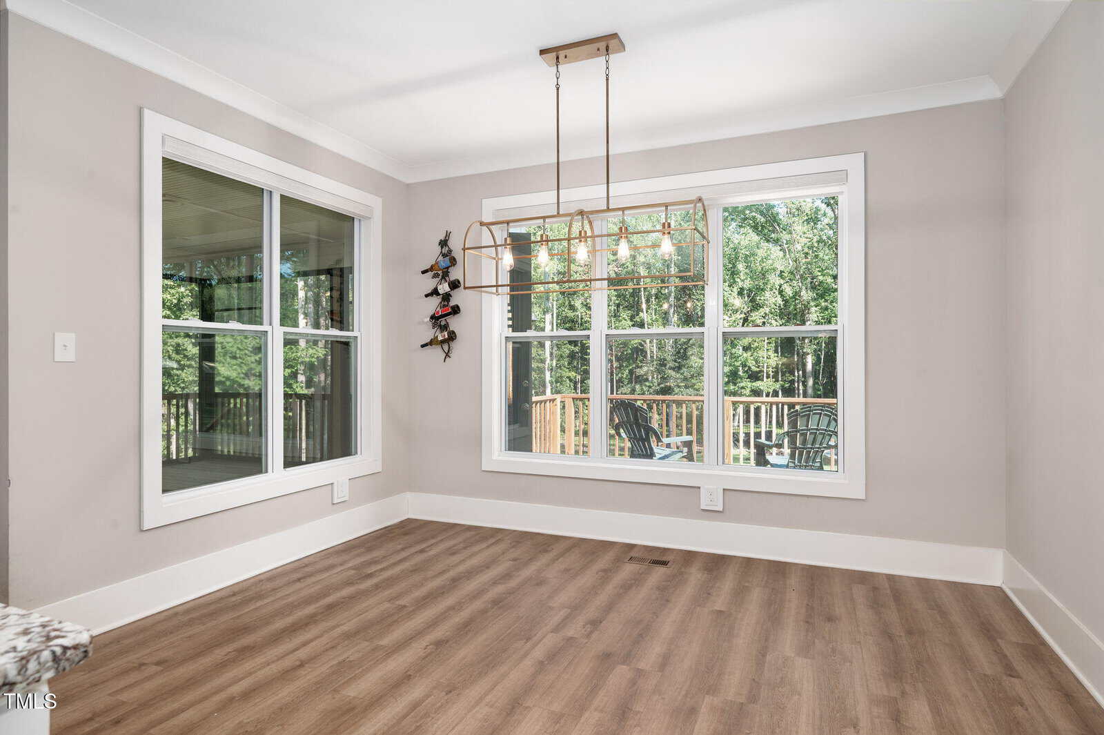 Bright dining area with large windows, modern chandelier, light wood flooring, neutral walls, and a wall-mounted wine rack. Outdoor deck with chairs and forest view visible through the windows.