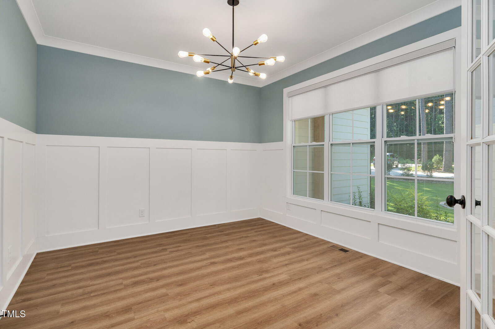 Bright, empty room with blue and white walls, large window with white blind, modern black chandelier, and wood flooring. Light streams in from outside through the window.