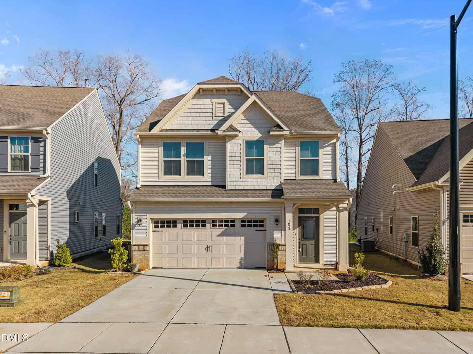 A two-story suburban house with light-colored siding, double garage, and a small covered front porch. There is a driveway and neatly maintained lawn, with neighboring houses on either side under a clear blue sky.