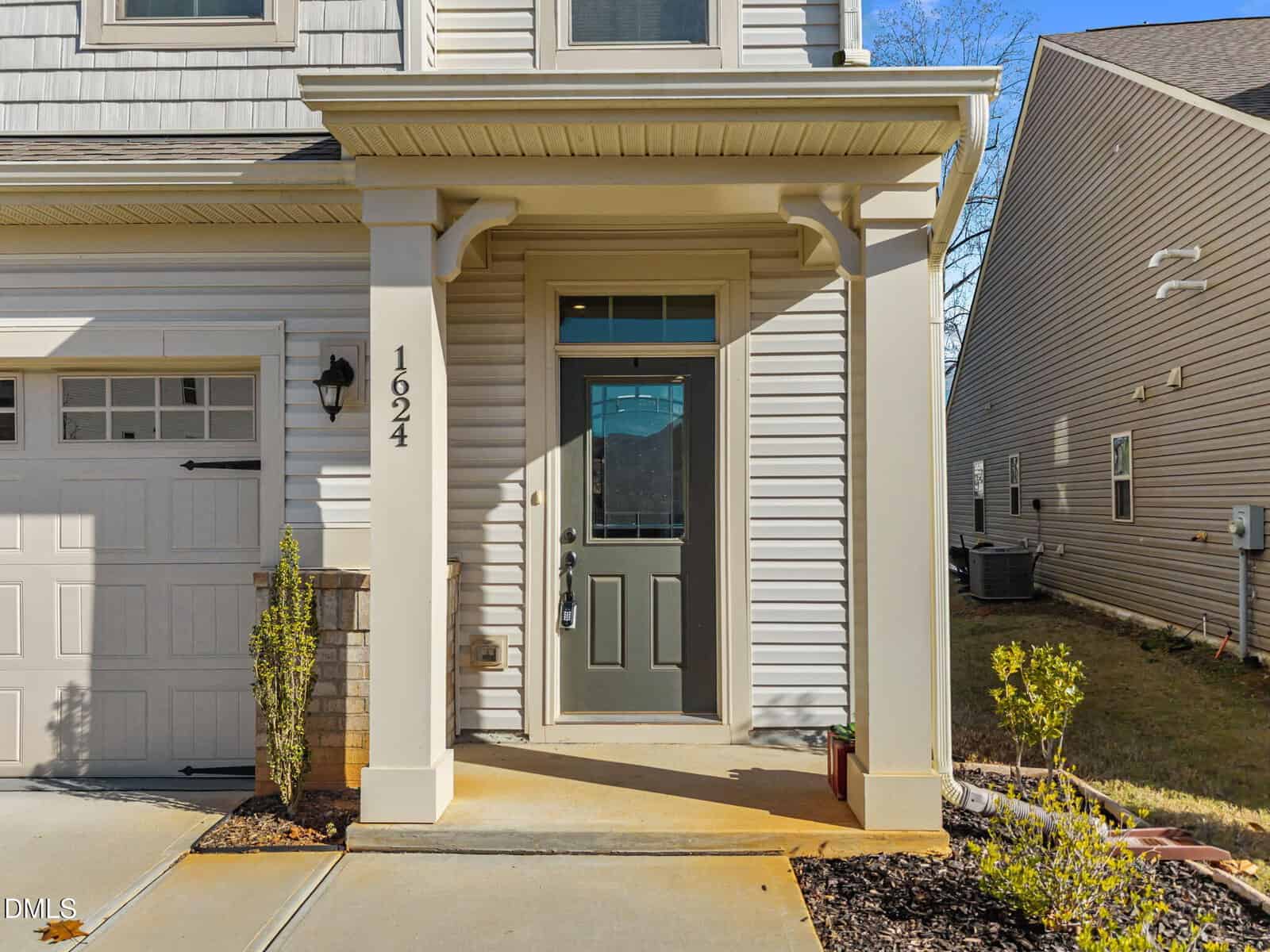 A modern house entryway with a green front door, white siding, and the number "1624" on a pillar. There are small shrubs and plants by the porch, with neighboring houses visible on each side.