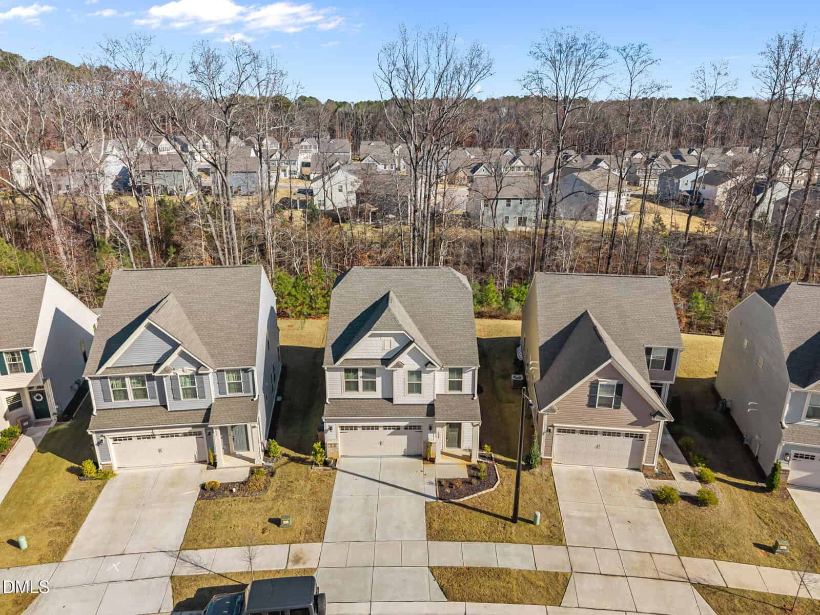 Aerial view of three modern, two-story suburban houses with driveways, light-colored exteriors, and neatly maintained lawns, set in a residential neighborhood bordered by trees.