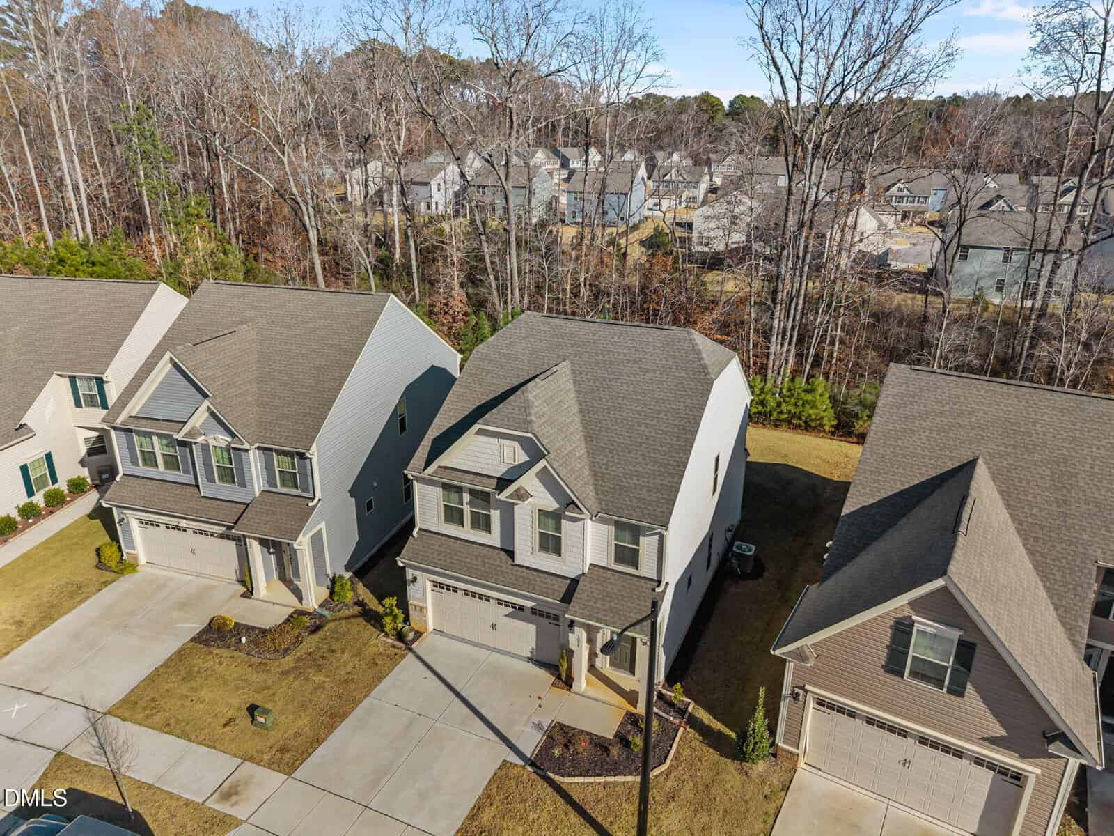 Aerial view of a row of modern two-story suburban houses with driveways, manicured lawns, and a wooded area in the background. The neighborhood appears quiet and residential.