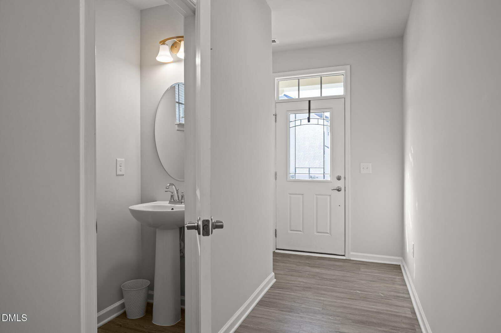 A narrow hallway with wood flooring, a white entry door with glass panels, and an open door revealing a small bathroom with a pedestal sink, round mirror, and wall-mounted light.