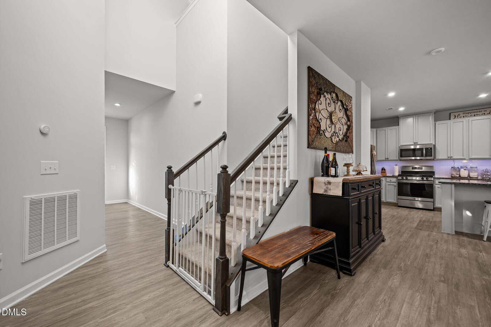 A modern entryway with wooden stairs featuring white spindles, a bench, and a sideboard beneath artwork. The space has light walls, wood flooring, and an open view to a kitchen with stainless steel appliances.