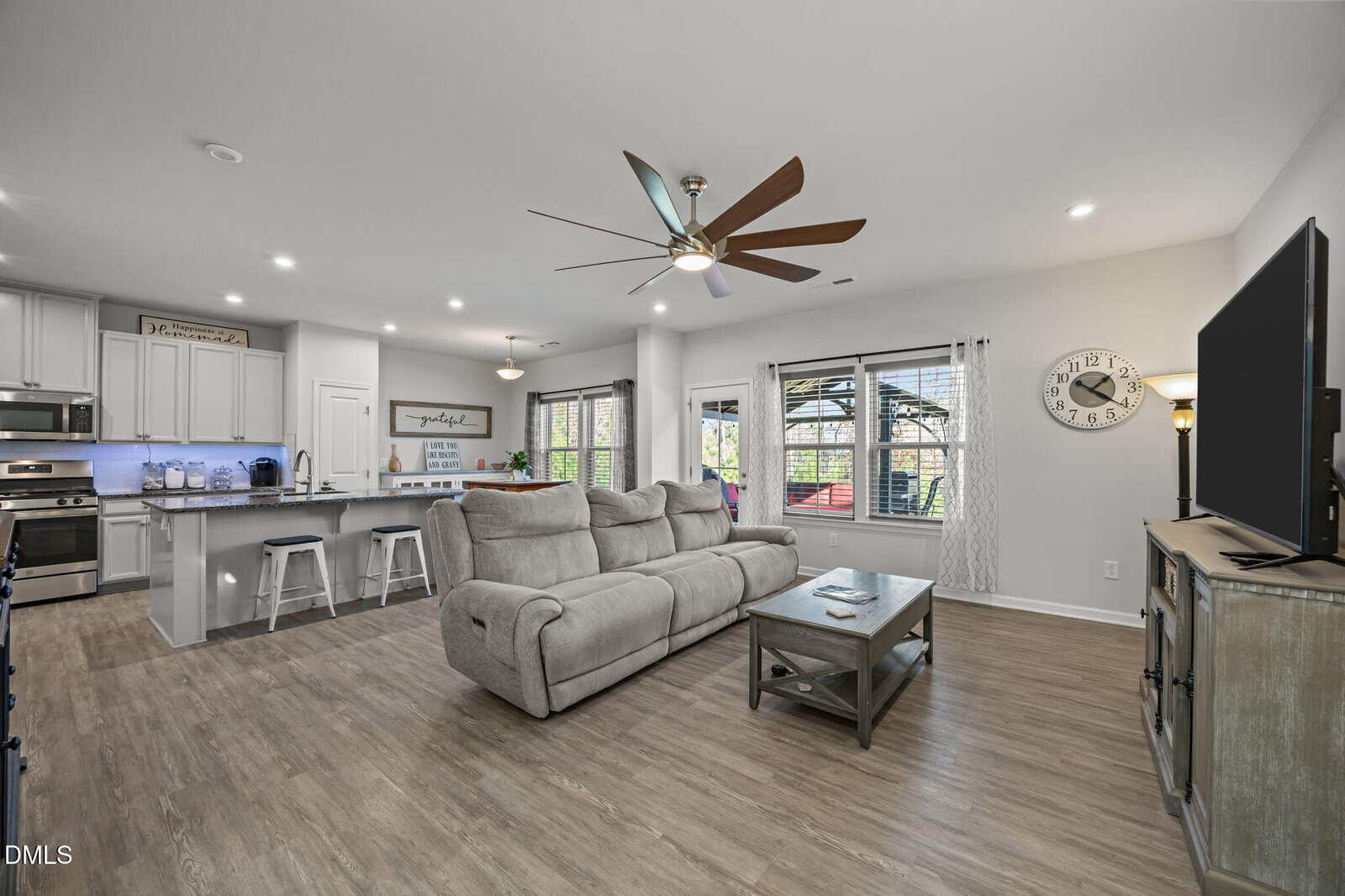 Open-concept living room and kitchen with gray sectional sofa, wooden coffee table, TV on a stand, modern ceiling fan, barstools at a kitchen island, stainless steel appliances, and large windows letting in natural light.