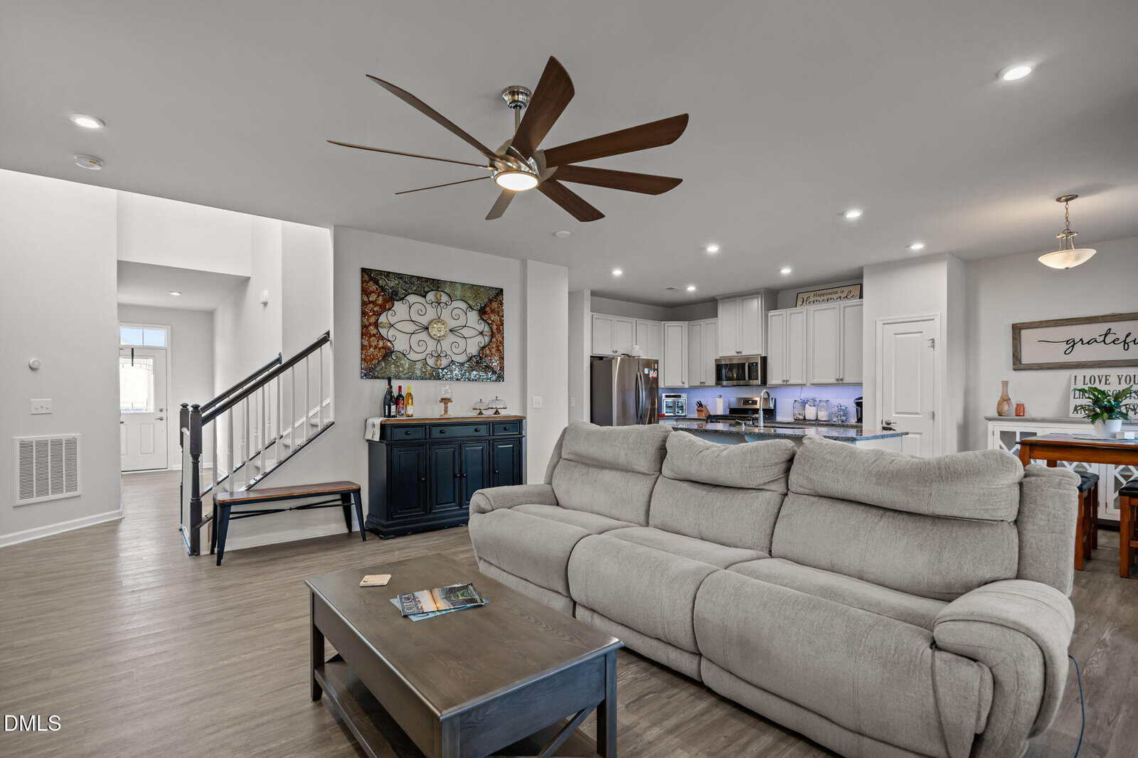 Modern open-concept living room with a large beige sofa, coffee table, and ceiling fan. The kitchen with white cabinets and stainless steel appliances is visible in the background, along with dining and entry areas.