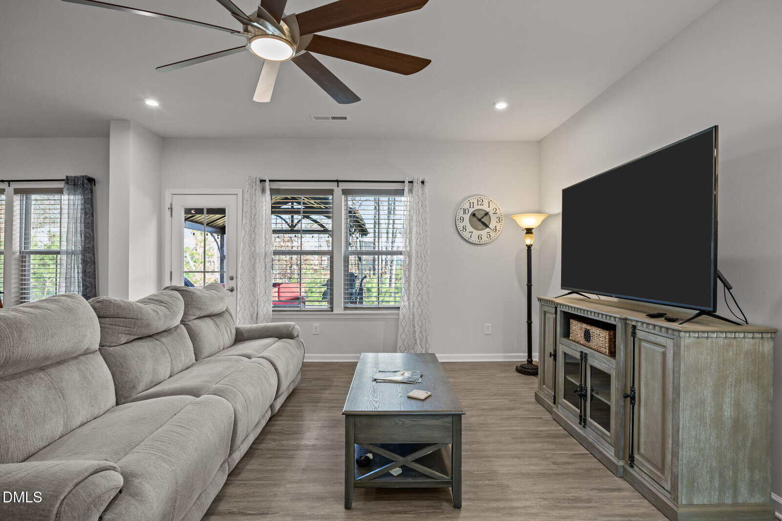 A modern living room with a large gray sectional sofa, a wooden coffee table, a flat-screen TV on a console, a floor lamp, and a wall clock. Large windows let in natural light and show greenery outside.