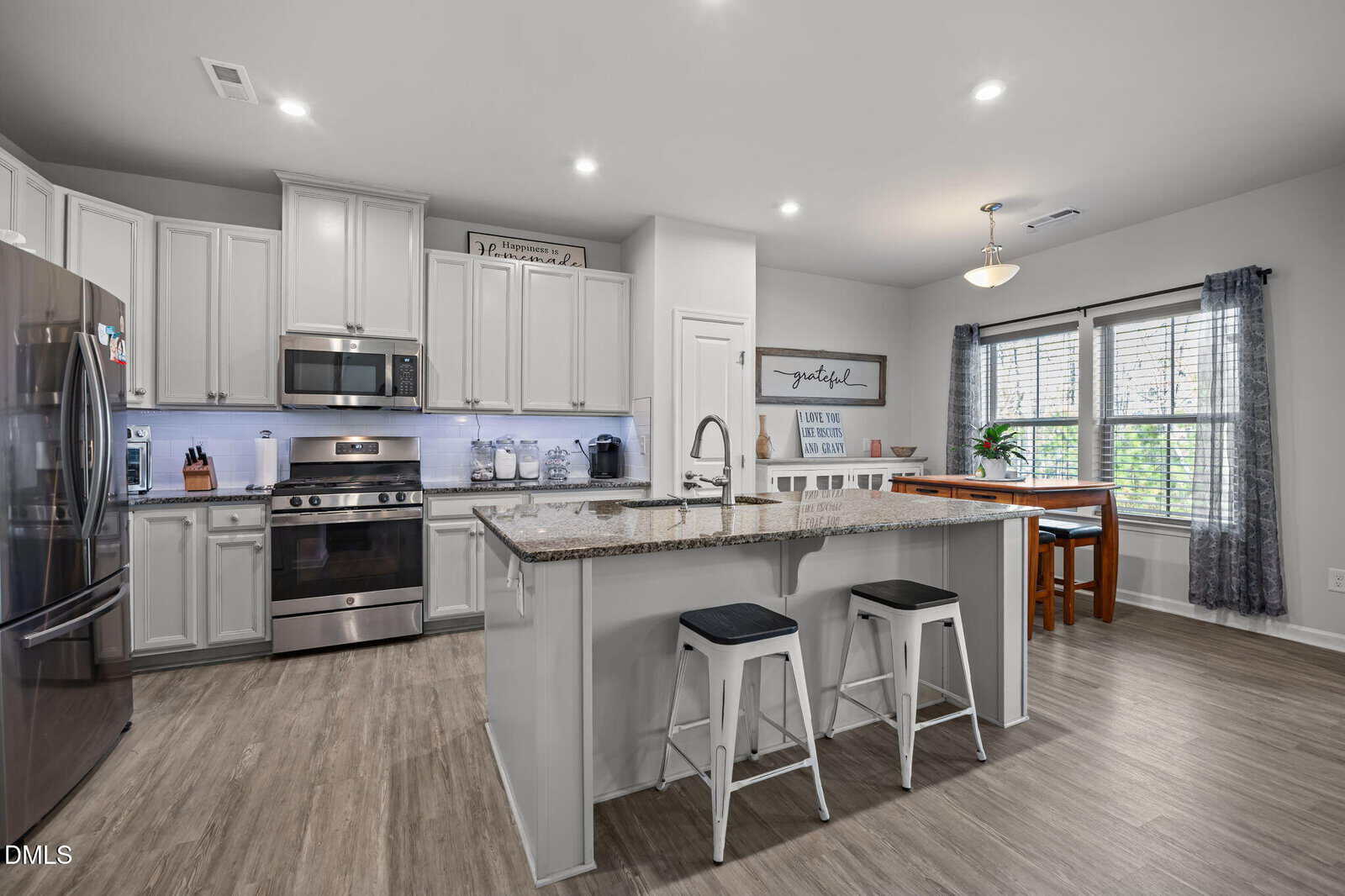 Modern kitchen with stainless steel appliances, white cabinets, a large island with a granite countertop and two stools, adjacent dining area with wooden table, and large windows with curtains.