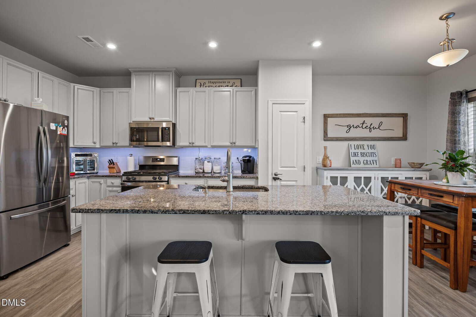 Modern kitchen with a large granite island, stainless steel appliances, white cabinets, and two barstools. A dining table is to the right, and decorative signs are displayed on the walls and above the cabinets.