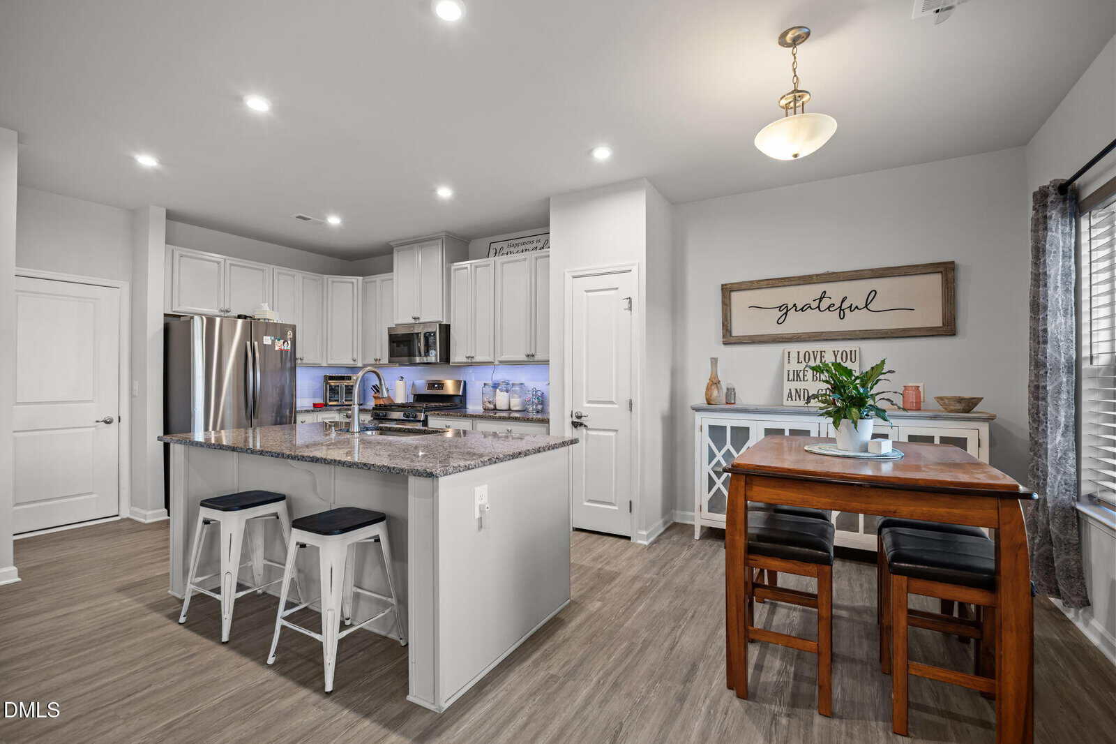 Modern kitchen with white cabinets, stainless steel appliances, island with two stools, dining table with two chairs, wood floors, and decorative signs on the wall. Warm, neutral tones and bright lighting throughout.