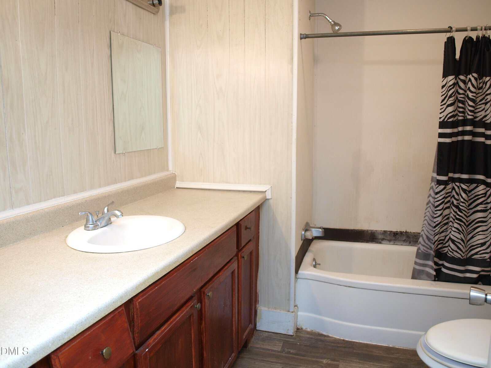A bathroom at 336 Vicksboro Road in Henderson features a single sink on a long beige countertop, wooden cabinets, a small mirror, a toilet, and a bathtub with zebra-striped shower curtain. The tub area shows visible wall stains.
