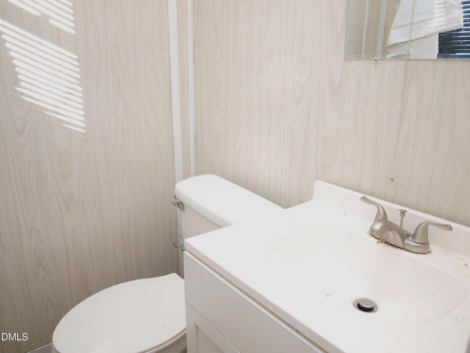 Small bathroom with a white toilet, a white sink vanity with a chrome faucet, light-colored wood panel walls, and a partial view of a mirror. Sunlight creates stripes on the wall.