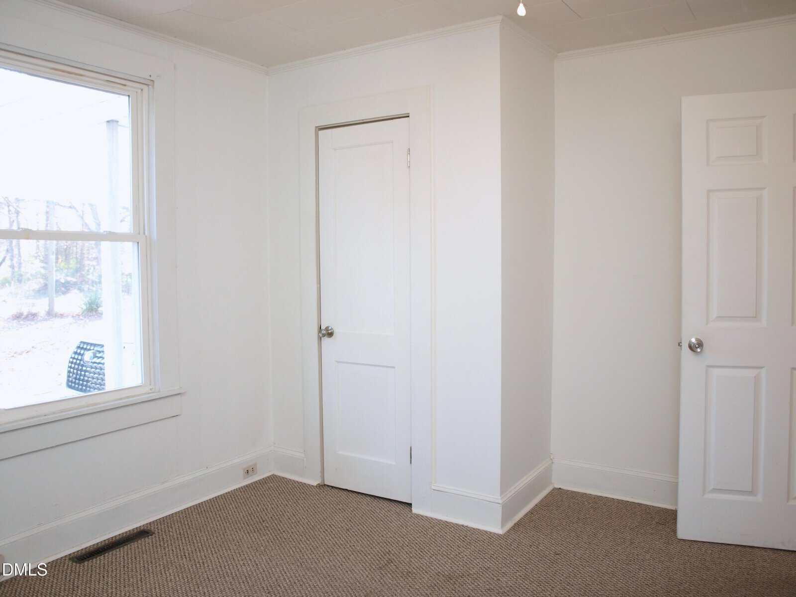 A small empty room with white walls, a beige carpet, a window on the left, a closed door in the center, and another door partially open on the right. Natural light comes through the window.