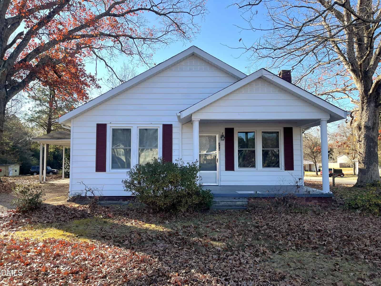 A single-story white house with a gabled roof, red shutters, and a small covered front porch. Leafless trees surround the house, and dry leaves cover the yard. A driveway and carport are visible to the left.