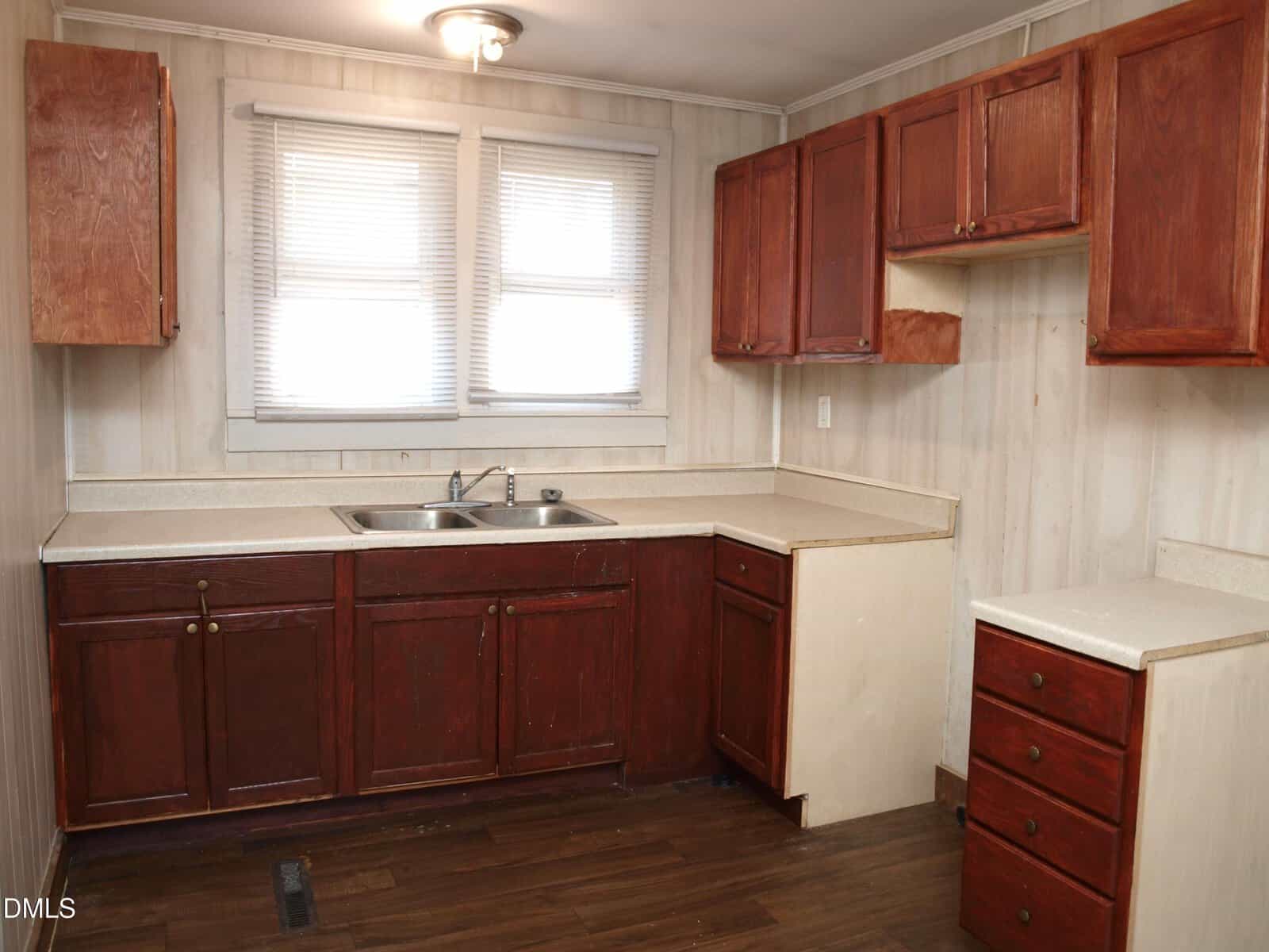 A small kitchen with dark wood lower cabinets, matching upper cabinets, light countertops, a stainless steel sink under two windows with blinds, and wood-patterned flooring. Walls are light-colored with a subtle pattern.