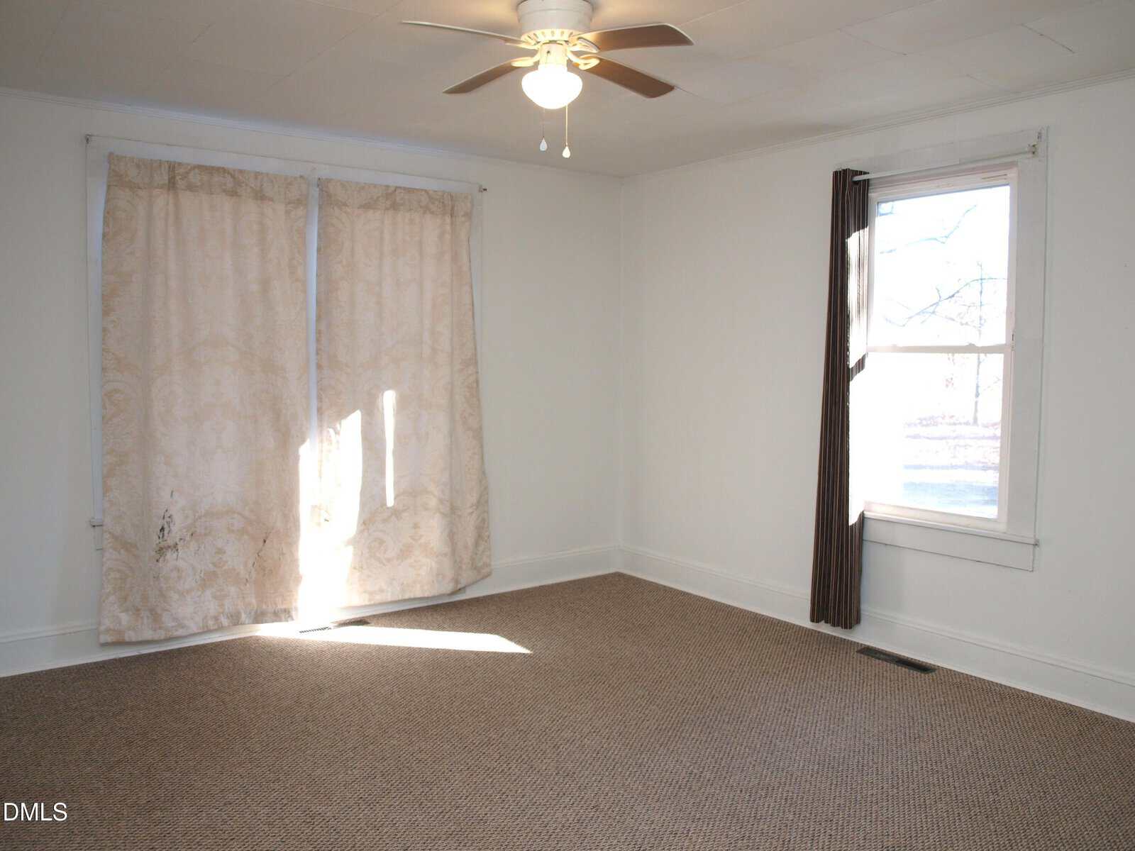 Empty room with beige carpet, white walls, a ceiling fan with light, a large window with brown curtains, and a set of covered double windows with patterned beige curtains. Sunlight shines through the curtain gap.