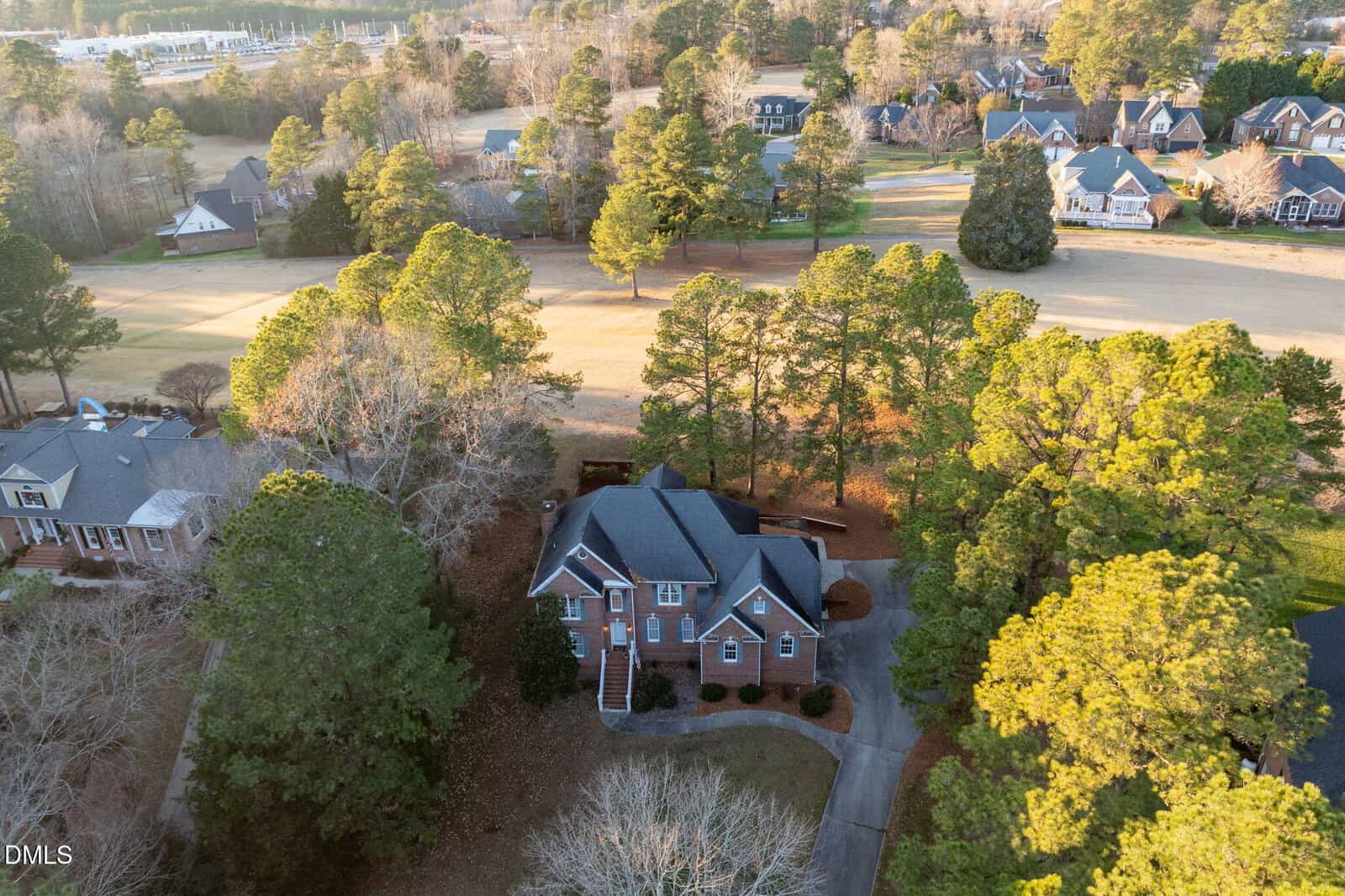 Aerial view of a two-story brick house surrounded by trees, with a curved driveway and a golf course or open grassy area behind it; neighboring houses and roads are visible in the background.