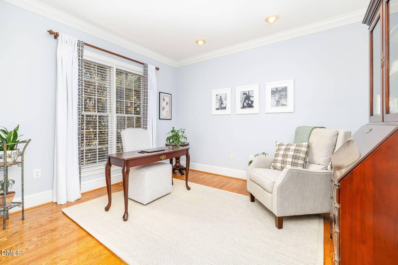 A bright home office with a wooden desk and gray chair near a large window, a light armchair with pillows, framed artwork on the wall, plants, wood floors, and a light rug.
