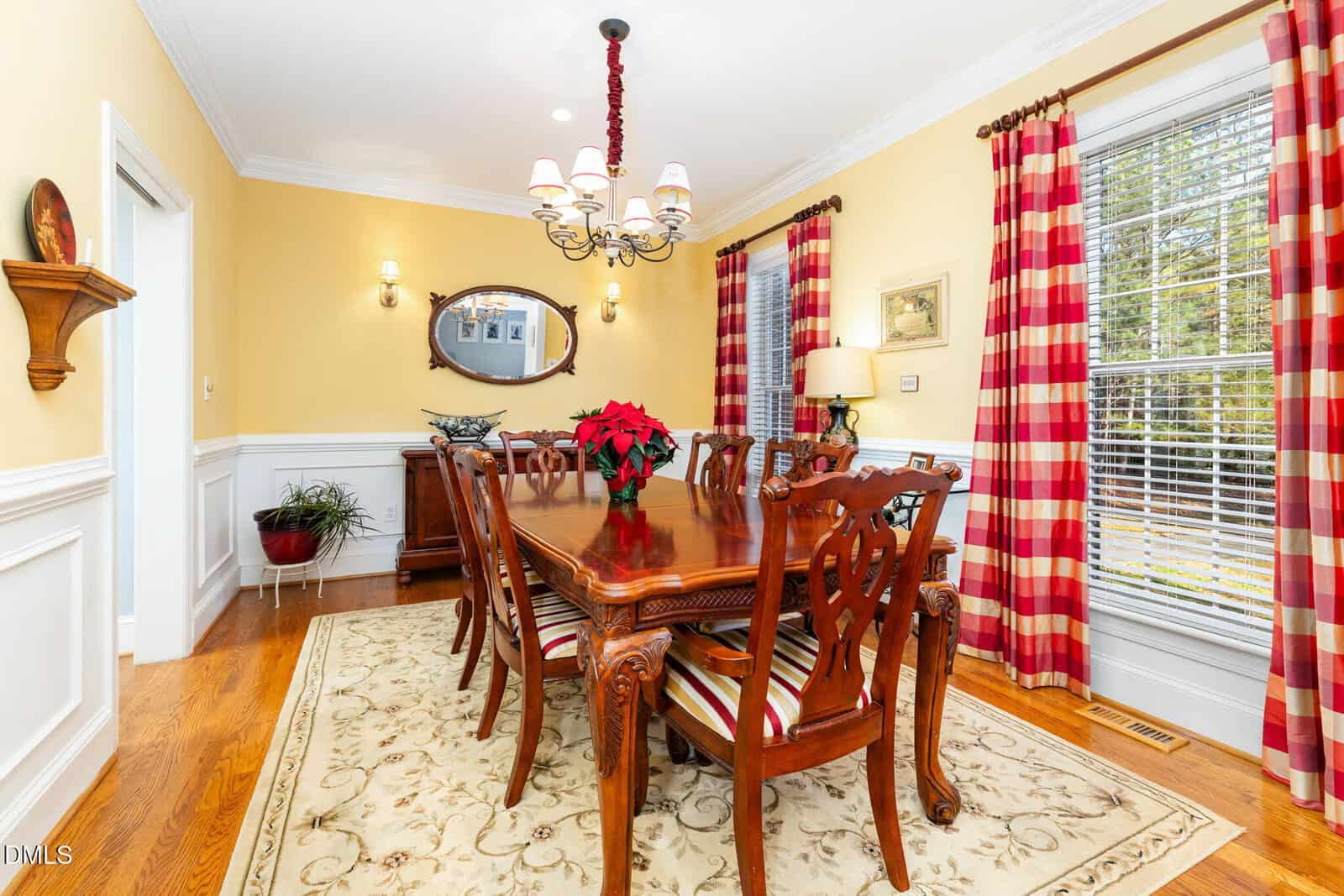 A formal dining room with a wooden table and eight chairs, a red poinsettia centerpiece, large windows with red plaid curtains, a chandelier, and a mirror above a sideboard against a yellow wall.