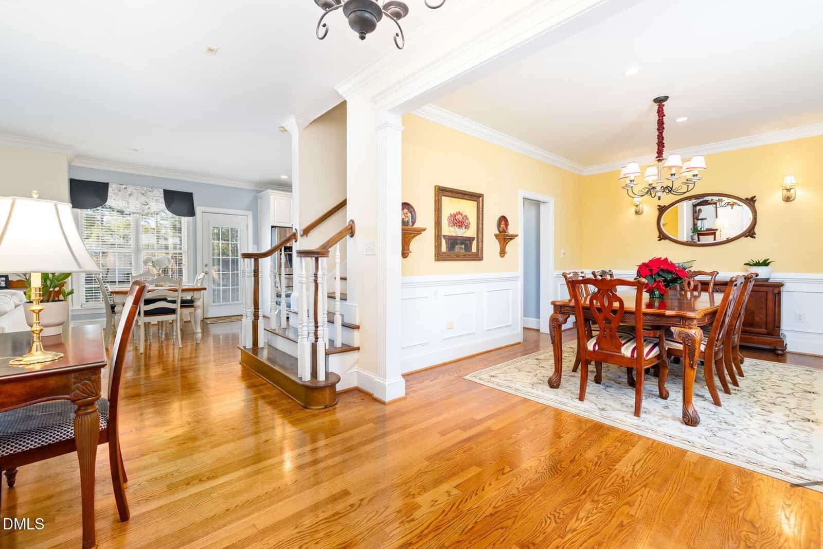 A spacious dining room with yellow walls and wood flooring, a large wooden dining table with chairs, chandelier, decorative mirror, and a side table. A staircase and sitting area are visible in the background.