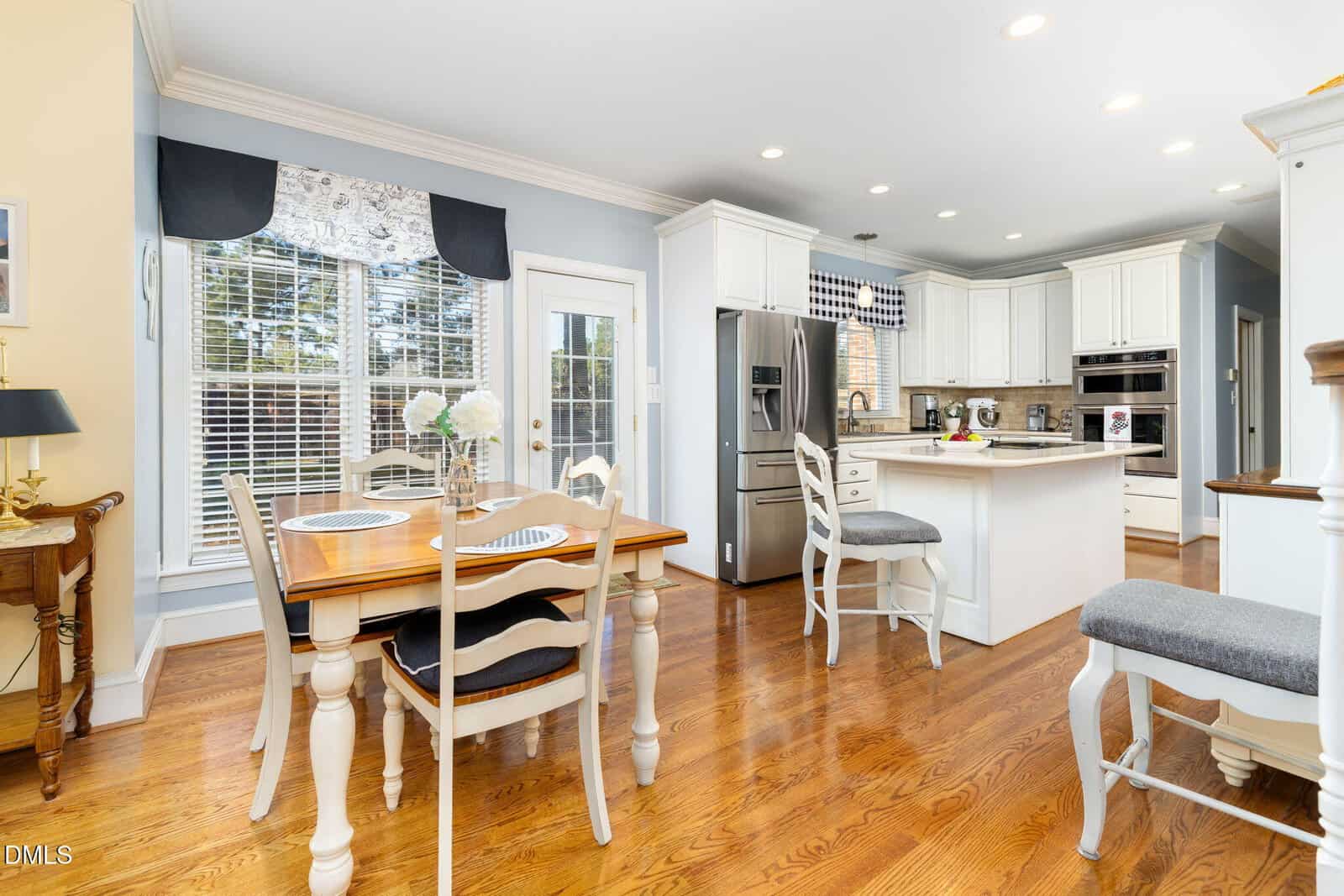 Bright, spacious kitchen with white cabinets, stainless steel appliances, a central island with stools, and a wooden dining table set for two next to large windows with blinds and valances. Hardwood floors throughout.