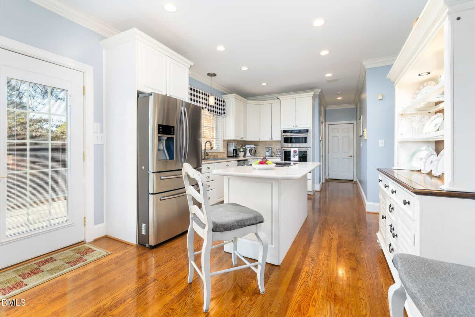 A bright, modern kitchen with white cabinets, stainless steel appliances, a center island with a gray cushioned chair, hardwood floors, and a hutch with white dishes on open shelves. Natural light enters through a glass door.