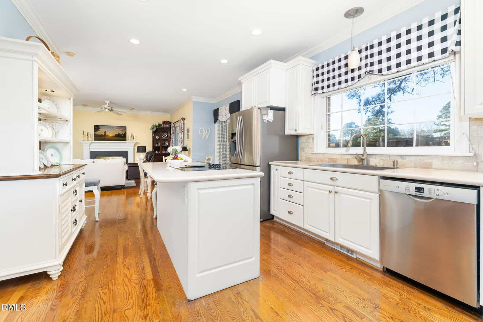 Bright kitchen with white cabinets, stainless steel appliances, a center island, and wooden floors. Window above the sink has a black-and-white checkered valance. Living room visible in the background.