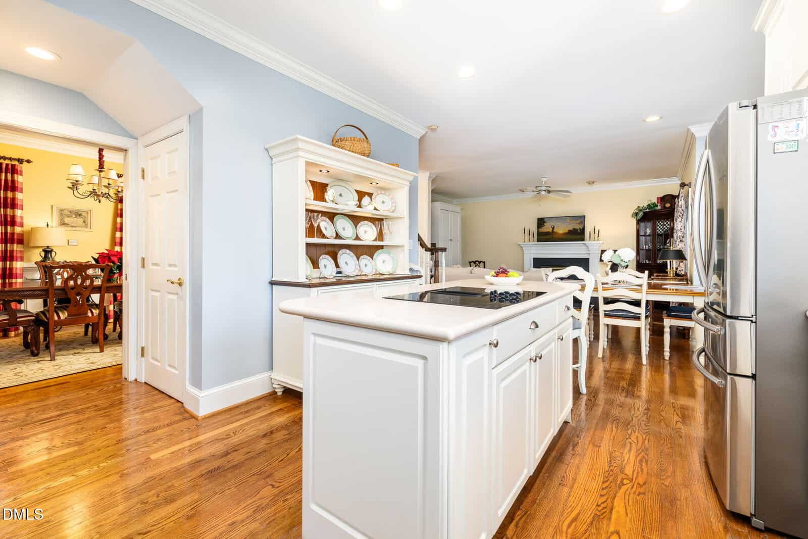 Bright kitchen with white cabinets, center island, stainless steel appliances, and hardwood floors. A dining area is visible in the background, and a separate formal dining room is seen to the left through an open doorway.
