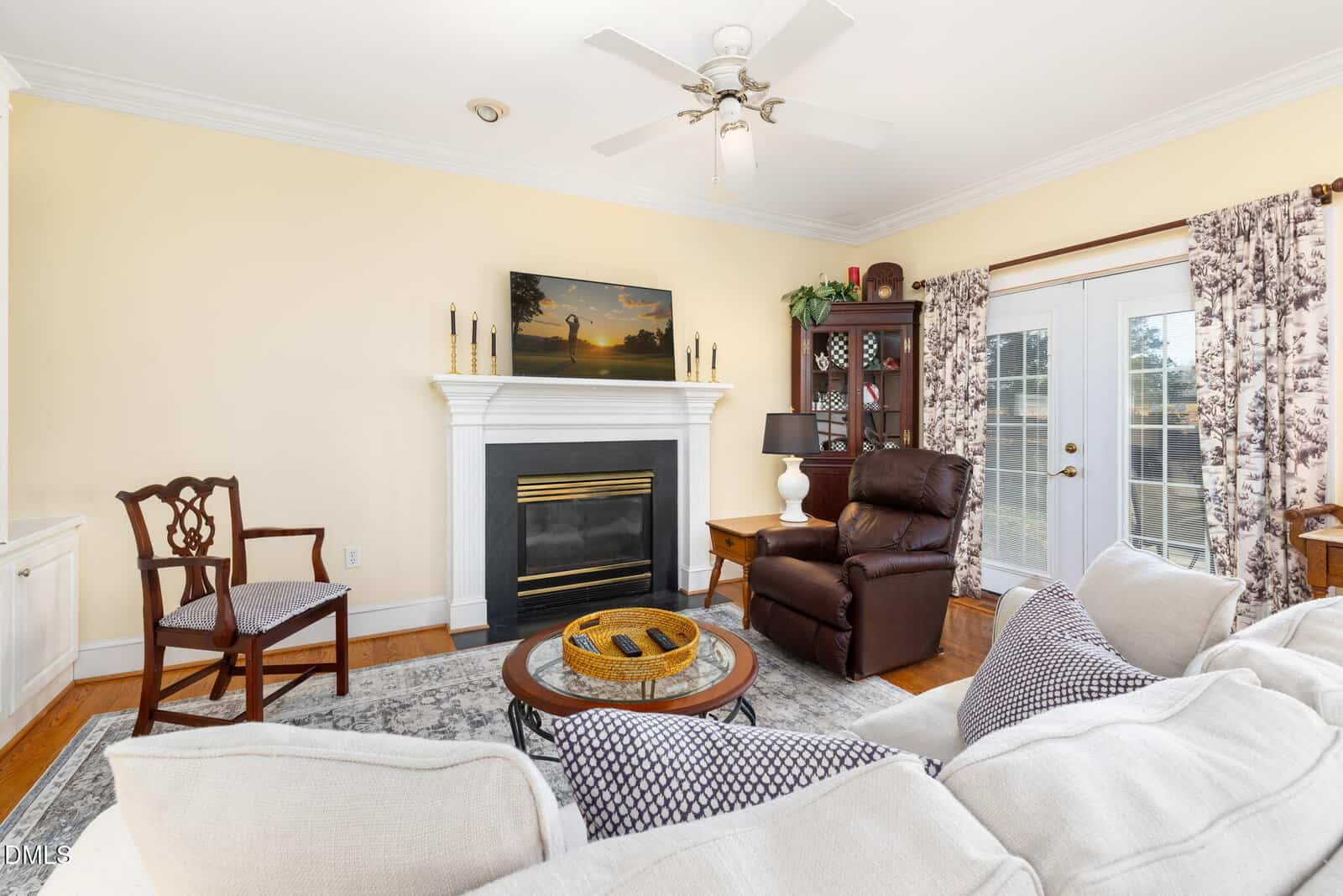 A living room with a white fireplace, TV above it, armchair, wooden chair, round coffee table, beige sofa, glass-door cabinet, lamp, and patterned curtains framing French doors. Walls are light yellow and floor is wood.