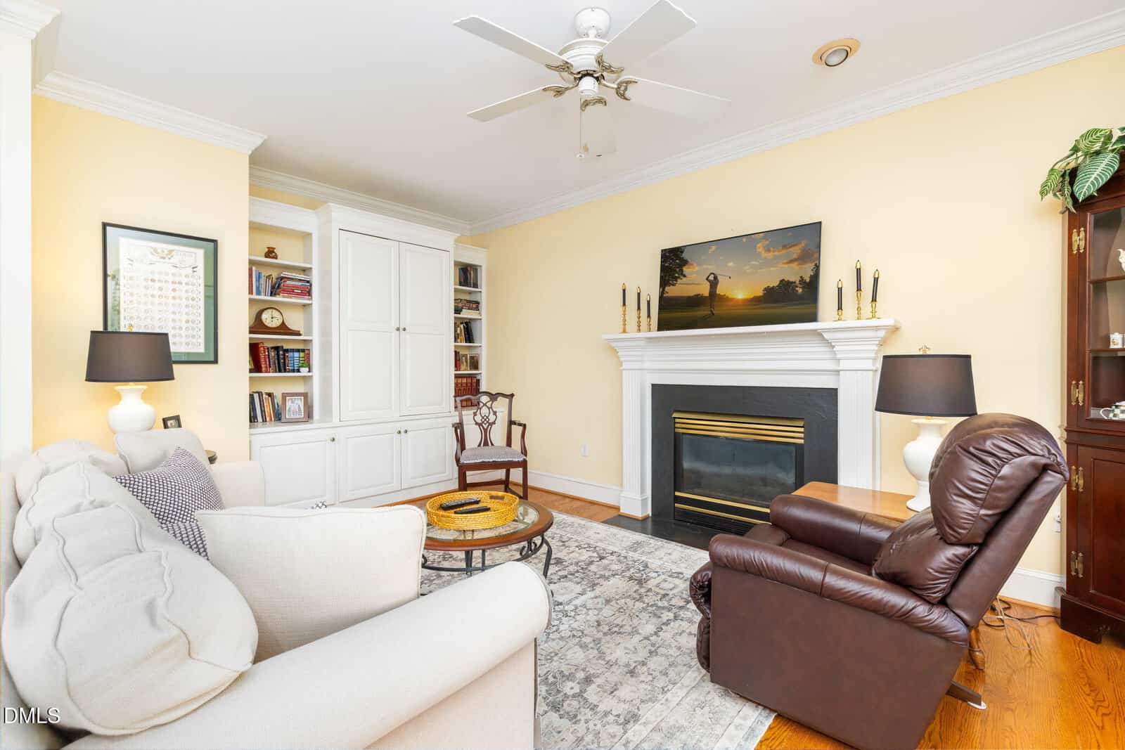 A living room with a beige couch, brown recliner, built-in white shelves, a fireplace with a TV above it, two black lamps, a rug, and hardwood floors. There is a ceiling fan and yellow walls.
