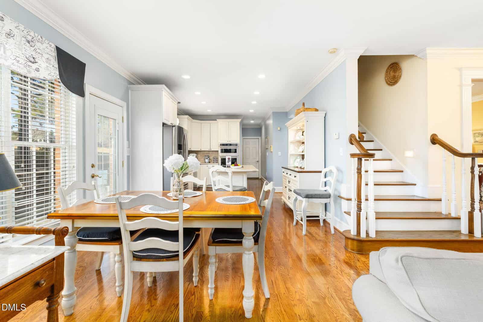 Bright kitchen and dining area with wooden floors, white cabinets, a wooden dining table set for four, and a stairway leading upstairs. Natural light fills the room through large windows and a glass door.
