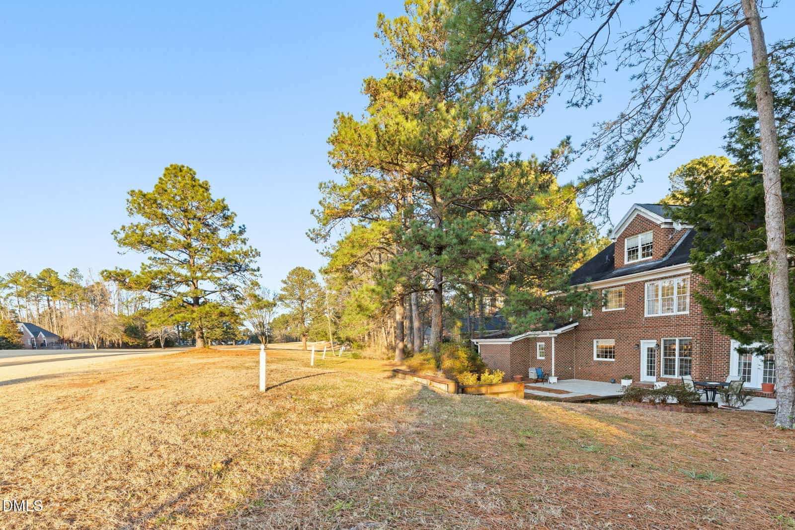 A two-story brick house with white-framed windows sits next to tall pine trees on a grassy, slightly sloped yard under a clear blue sky. A paved driveway is partially visible on the left.