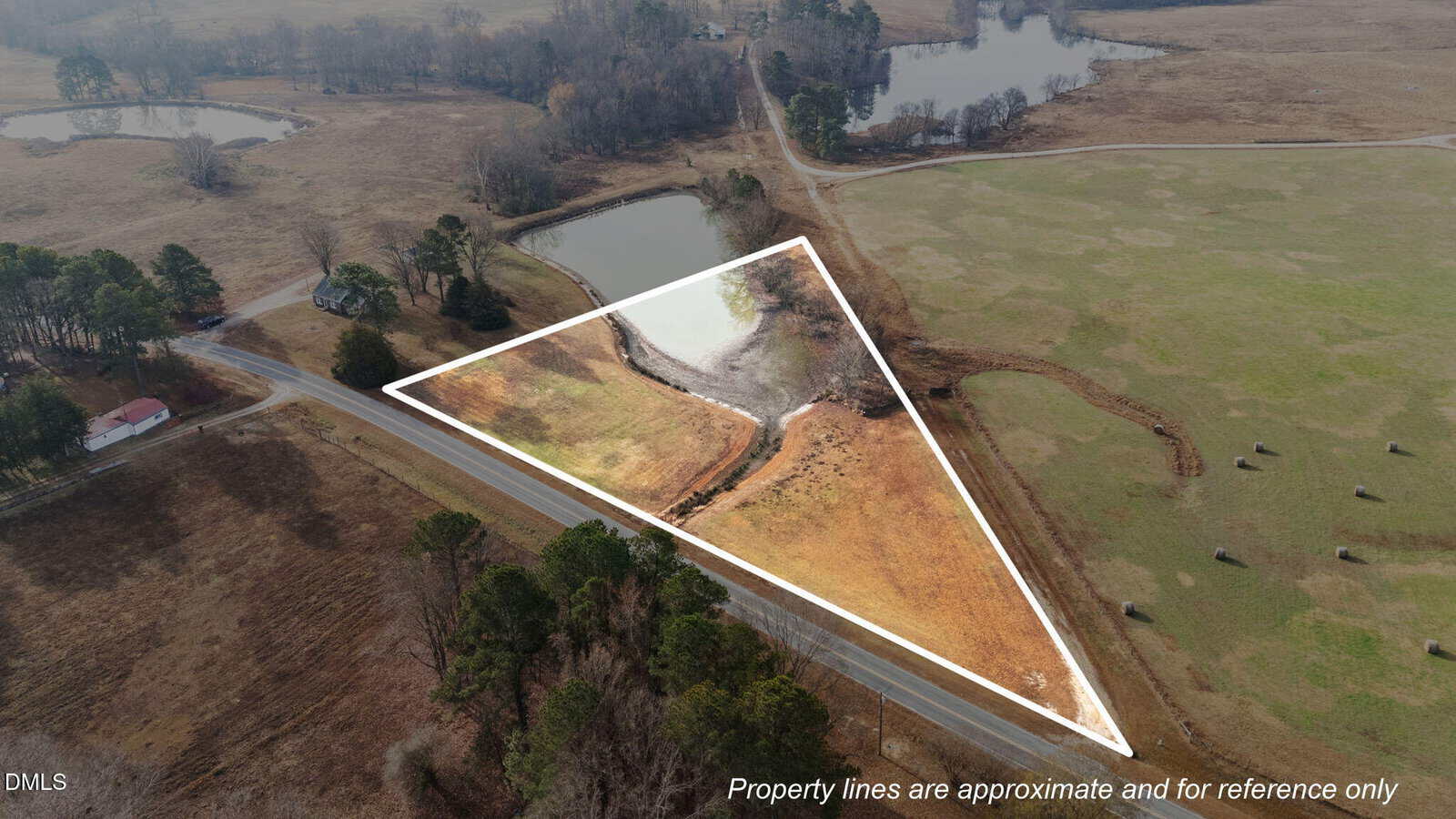 Aerial view of a triangular plot of land outlined in white, bordered by a road, a pond, and open fields, with trees, a house, and hay bales nearby; text notes property lines are approximate.