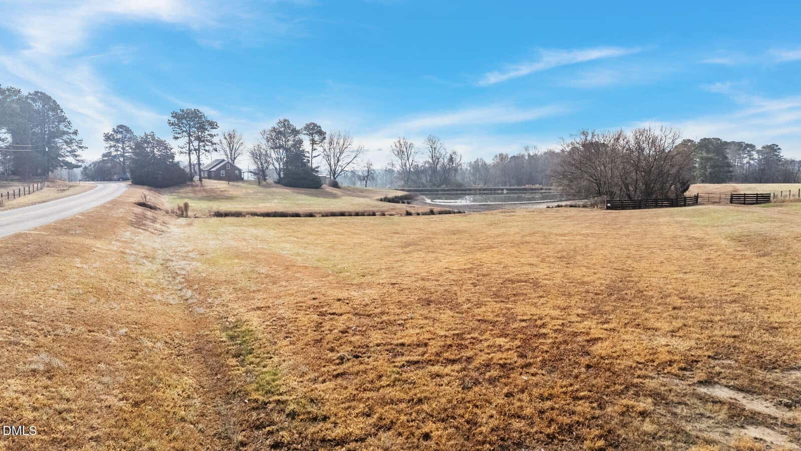 A wide, open field with dry grass, bordered by a road on the left and a pond in the background. Trees and a house are visible in the distance under a clear blue sky.