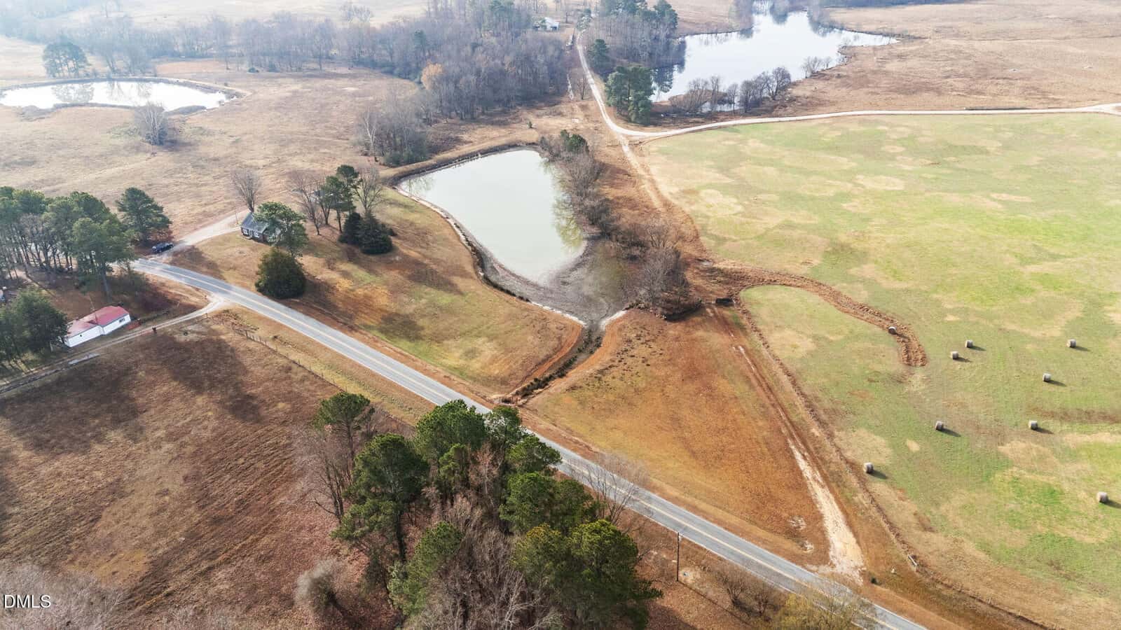 Aerial view of a rural landscape with a paved road, scattered trees, ponds, fields with hay bales, and a few houses. The land is mostly open and brown with patches of green grass.
