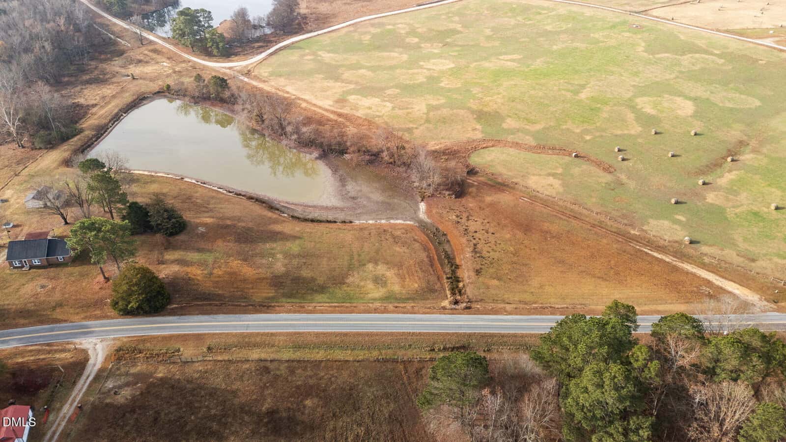 Aerial view of a rural landscape with a small pond, a winding creek, grassy fields, round hay bales, a country road, and a house surrounded by trees.