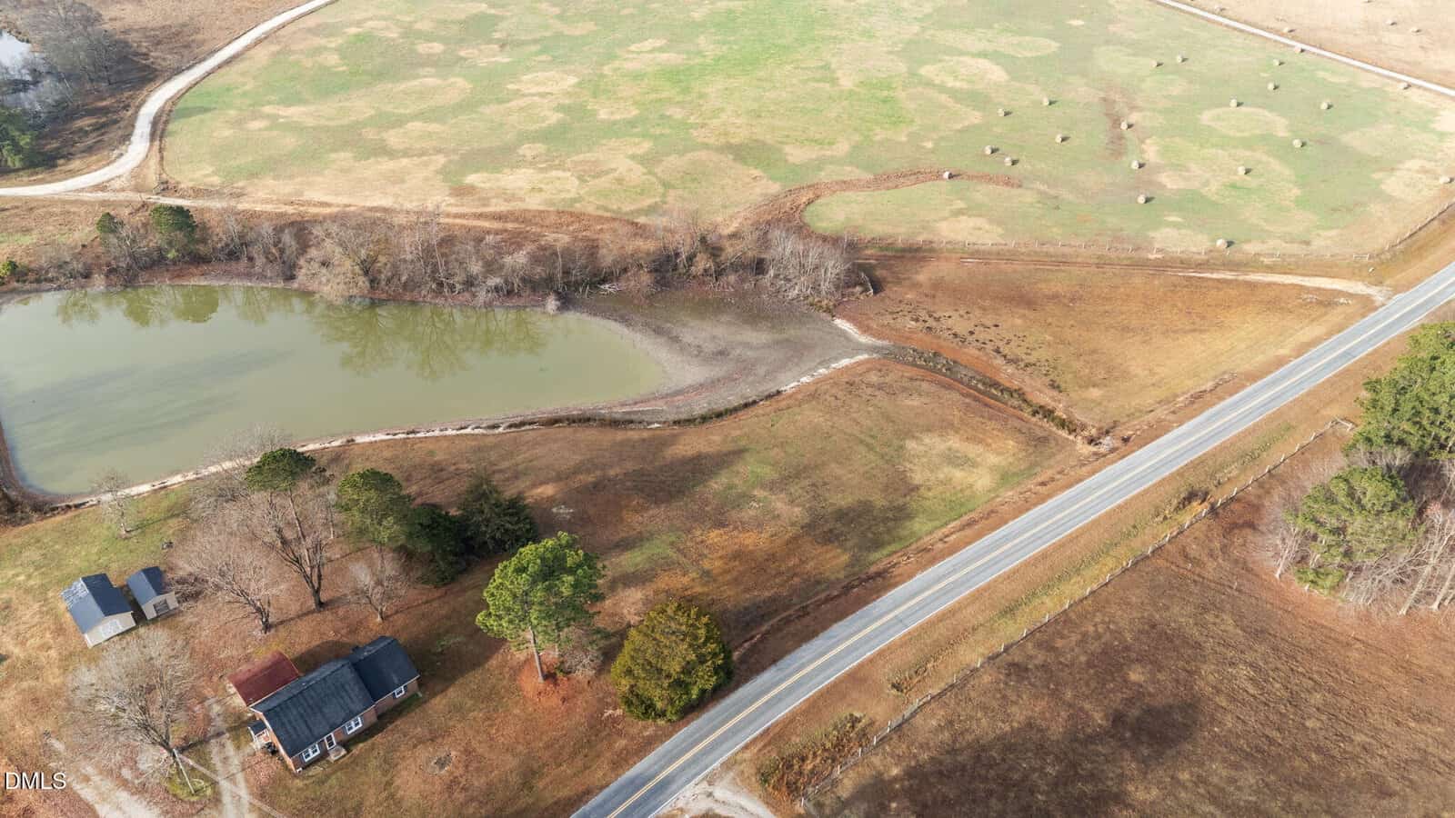 Aerial view of a rural landscape featuring a house, a pond, open fields with hay bales, scattered trees, and a road running along the edge of the property.