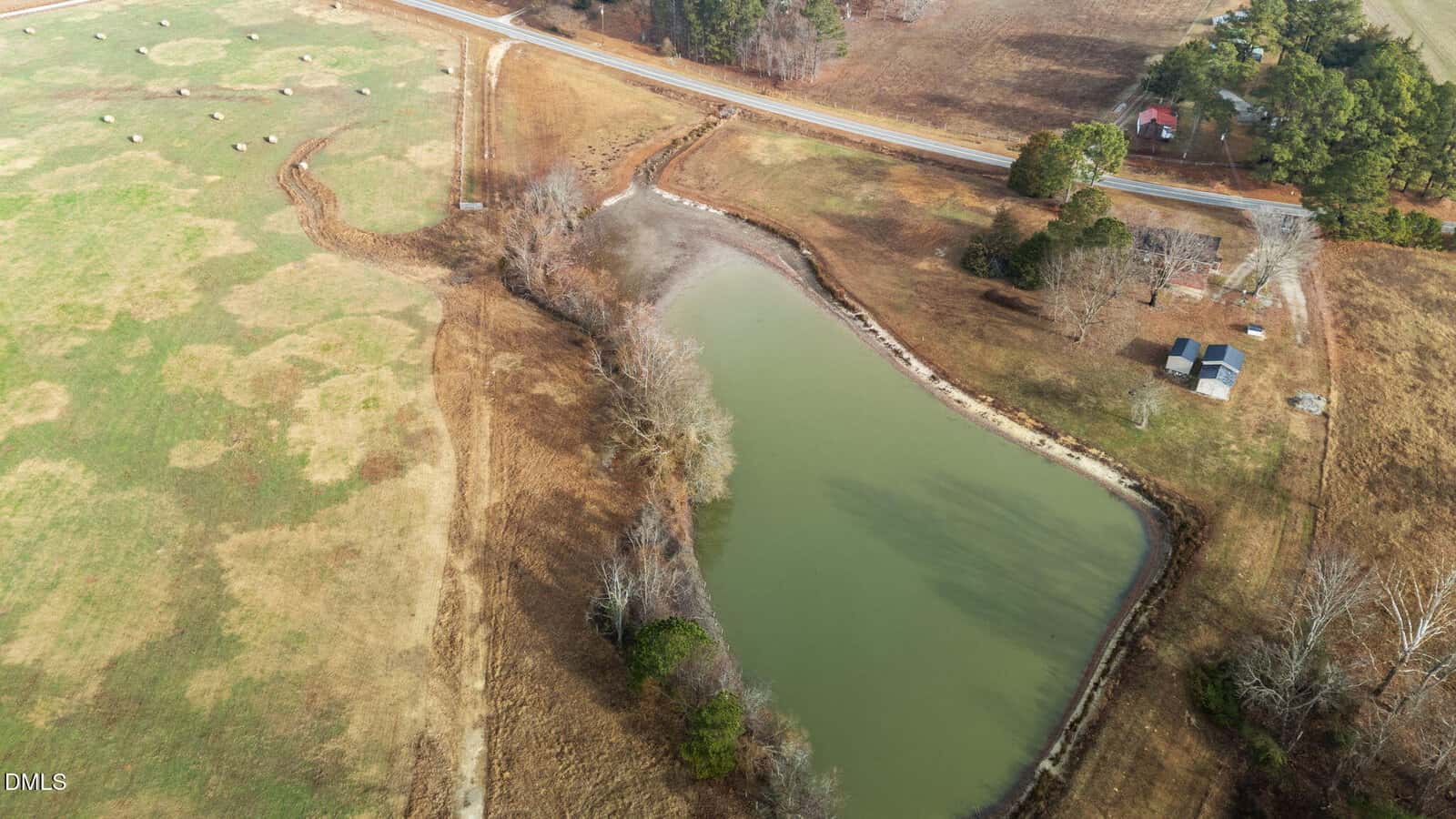 Aerial view of a small pond surrounded by brown grass and trees, with a road and scattered houses nearby and a green pasture with hay bales to the left.