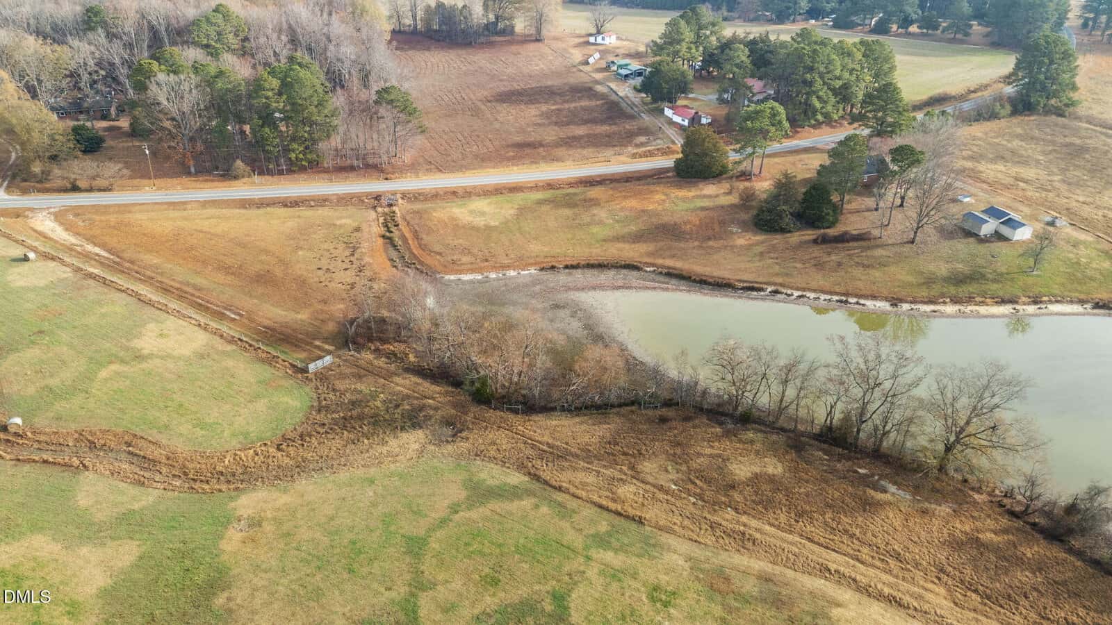 Aerial view of a rural landscape featuring a small pond, grassy fields, a road running through the scene, scattered trees, and several buildings near the roadside.