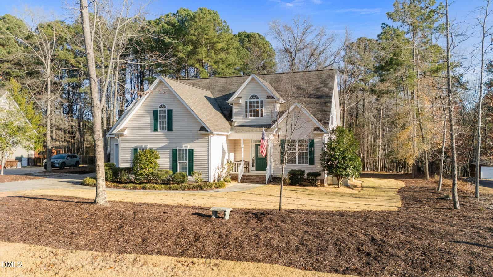 A two-story house with gray siding, white trim, green shutters, and an American flag by the front porch. The yard has mulch, trees, and grass, with a wooded area in the background under a clear blue sky.
