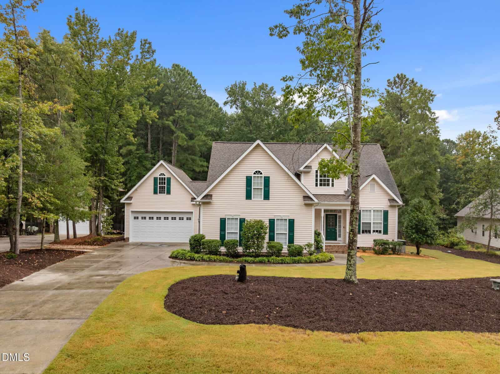 Nestled at 155 Oak Leaf Trail in Youngsville, this two-story beige house features a gray roof, an attached two-car garage, and an inviting front porch. The home is surrounded by mature trees and a landscaped lawn, with a mulched area and concrete driveway in front. The sky is clear and blue, offering a picturesque setting for this charming Youngsville residence.