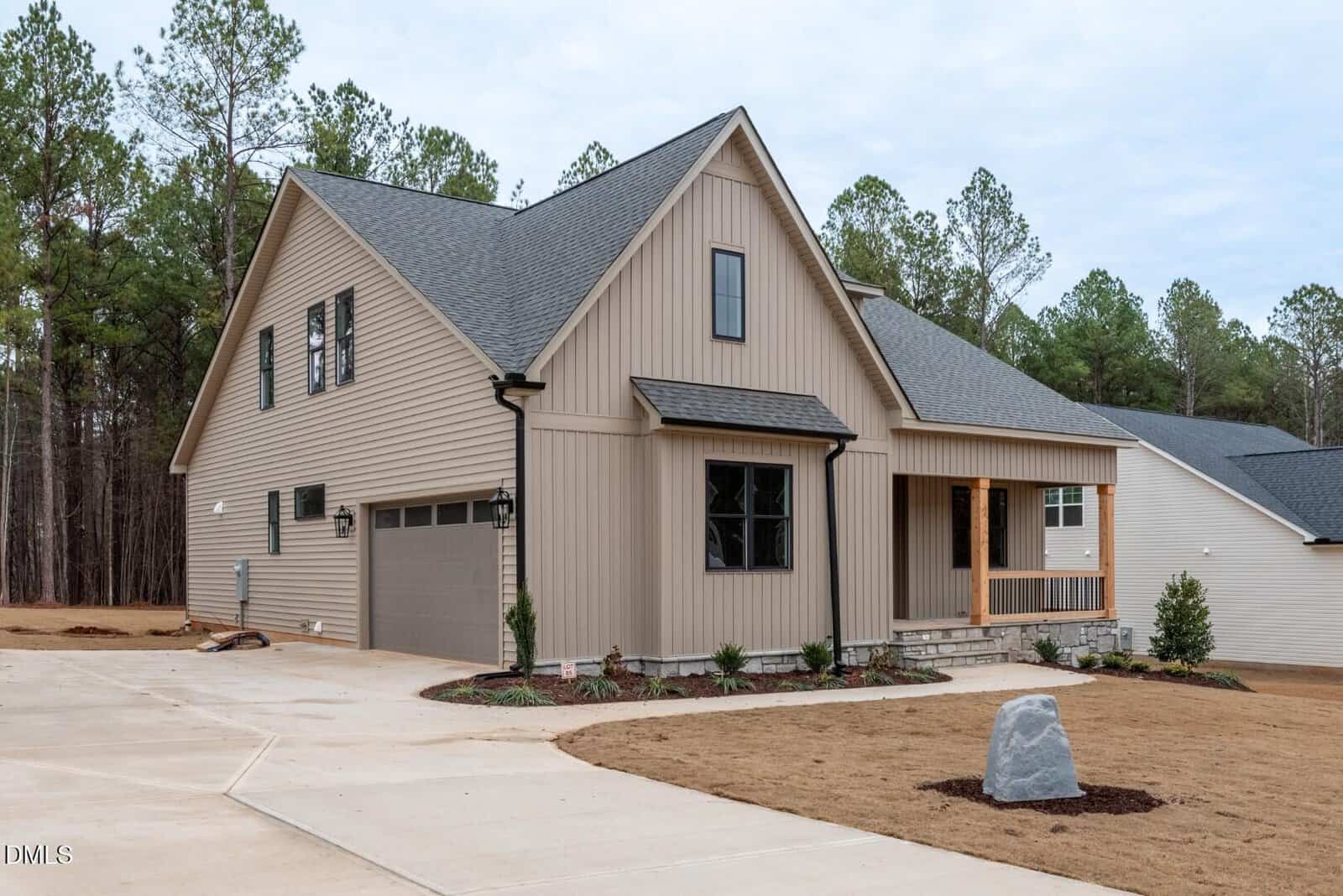A modern beige house with dark roofing and a two-car garage, surrounded by minimal landscaping and set on a corner lot with a concrete driveway. Pine trees line the background under an overcast sky.