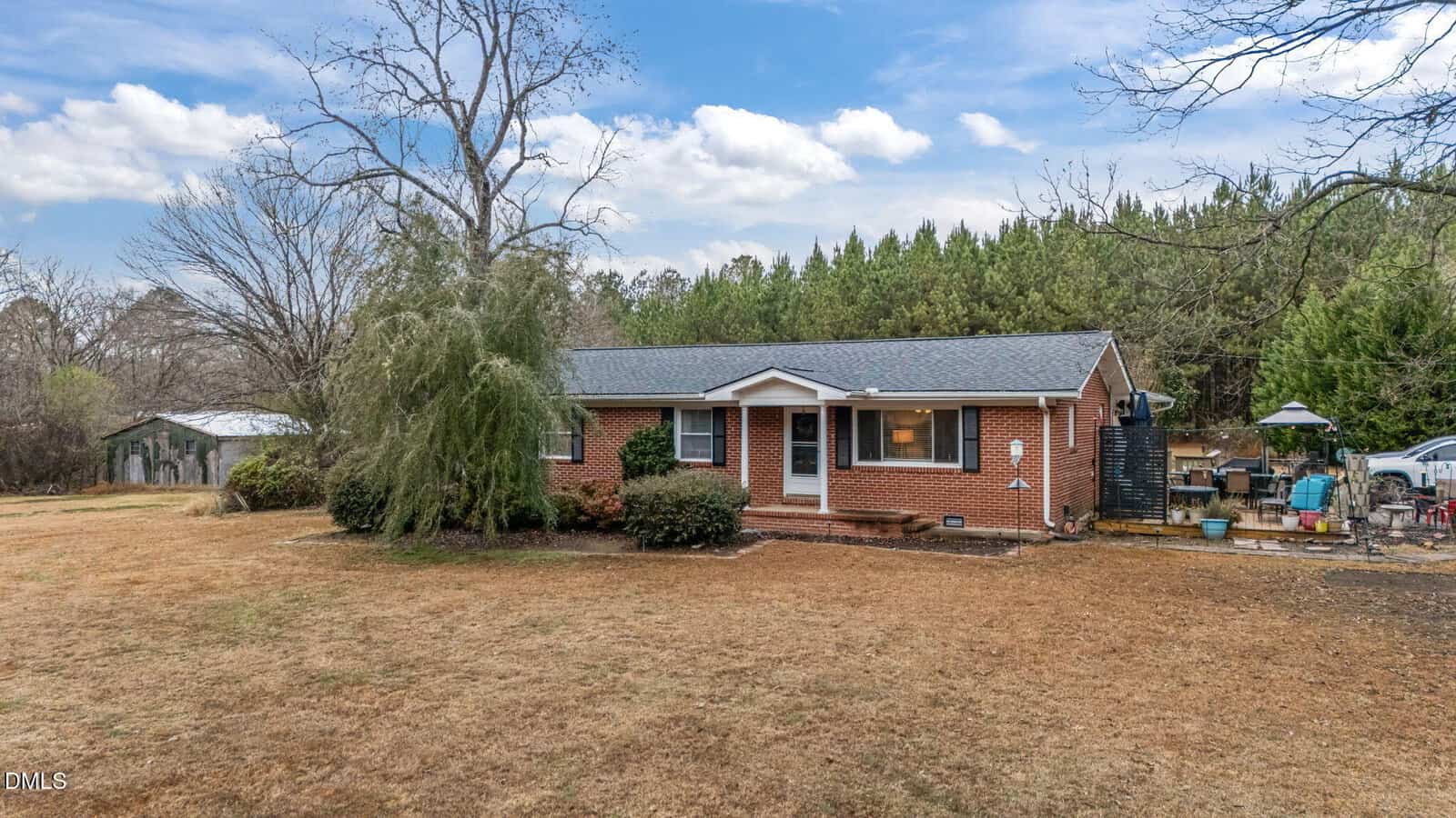 A single-story brick house with a gray shingle roof sits on a dry, grassy yard at 4553 Old NC 75, Oxford. There are a few bushes near the home, trees in the background, outdoor furniture on a patio, and a small outbuilding to the left.