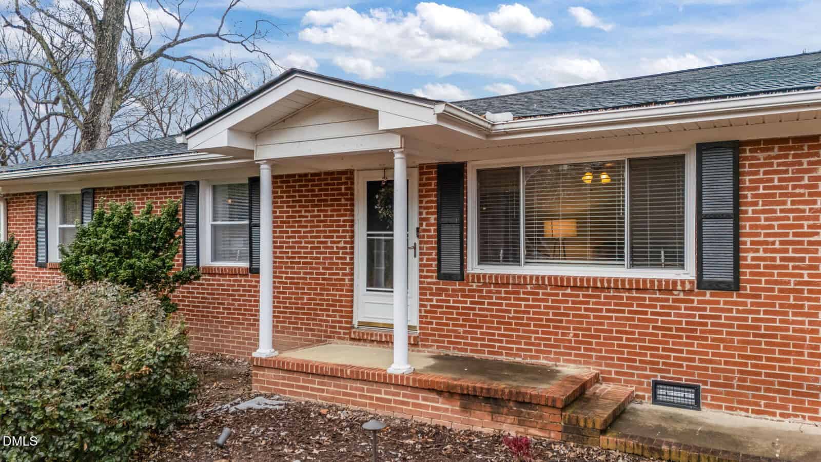 A brick house with white trim and columns, featuring a covered entryway, a white door with glass inset, two windows with blinds, and a small step leading to the entrance. Shrubs and bare soil are in the foreground.