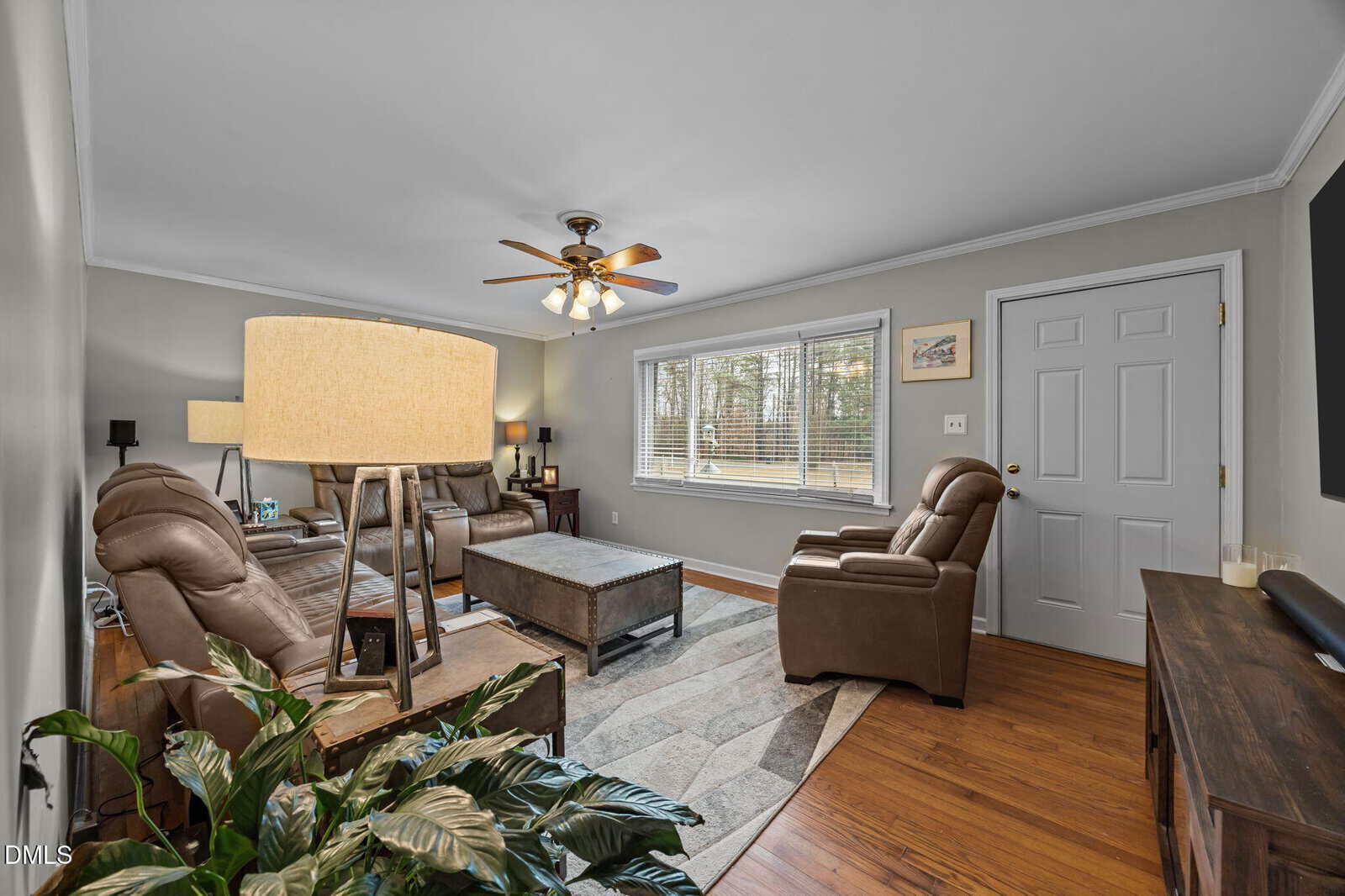 A living room with brown leather recliners, a wooden coffee table, a large area rug, ceiling fan with lights, a window with blinds, a door, and hardwood floors. A floor lamp and green plant are in the foreground.