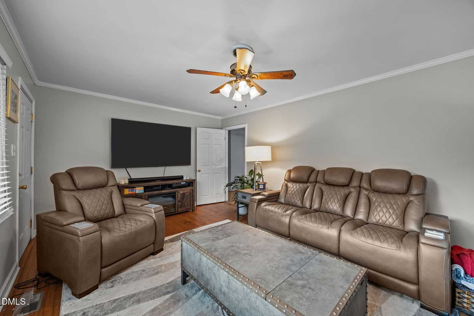 A living room with a brown leather recliner and matching sofa, a large ottoman-style coffee table, a TV on a wooden console, a floor lamp, a ceiling fan, and light gray walls with white trim.