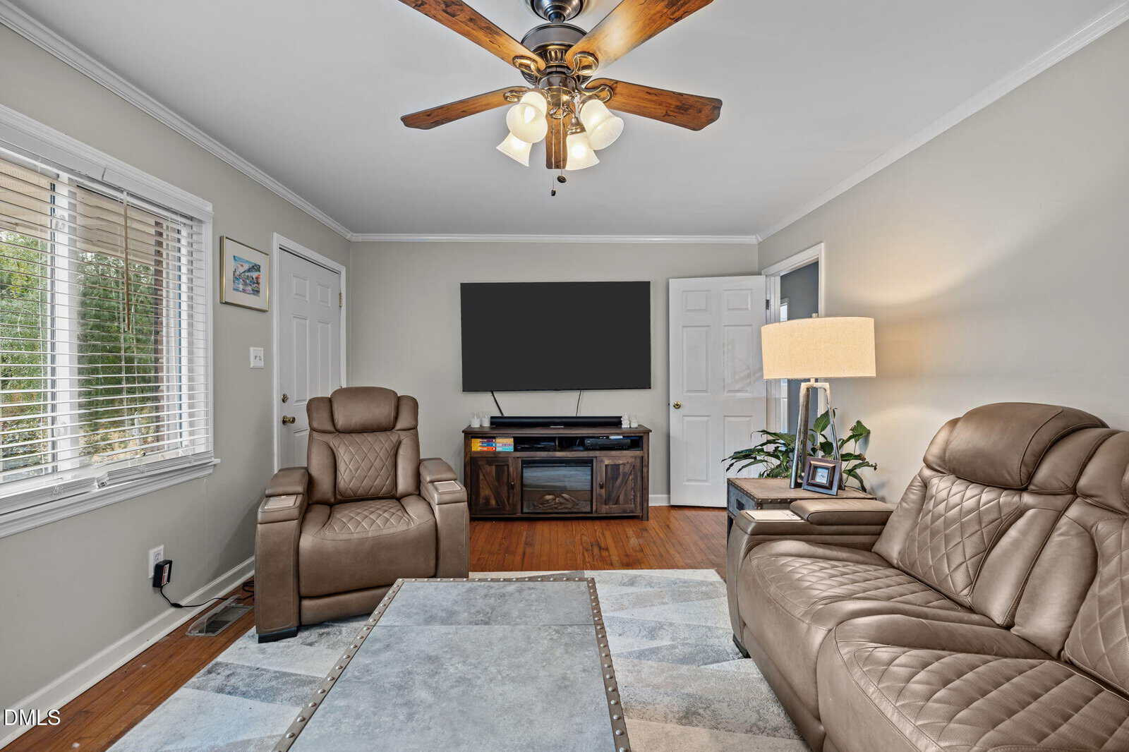 A living room with two brown leather chairs, a brown sofa, a ceiling fan with lights, a large TV on a stand, a table, a lamp, and two windows with blinds. The floor is hardwood and walls are light gray.