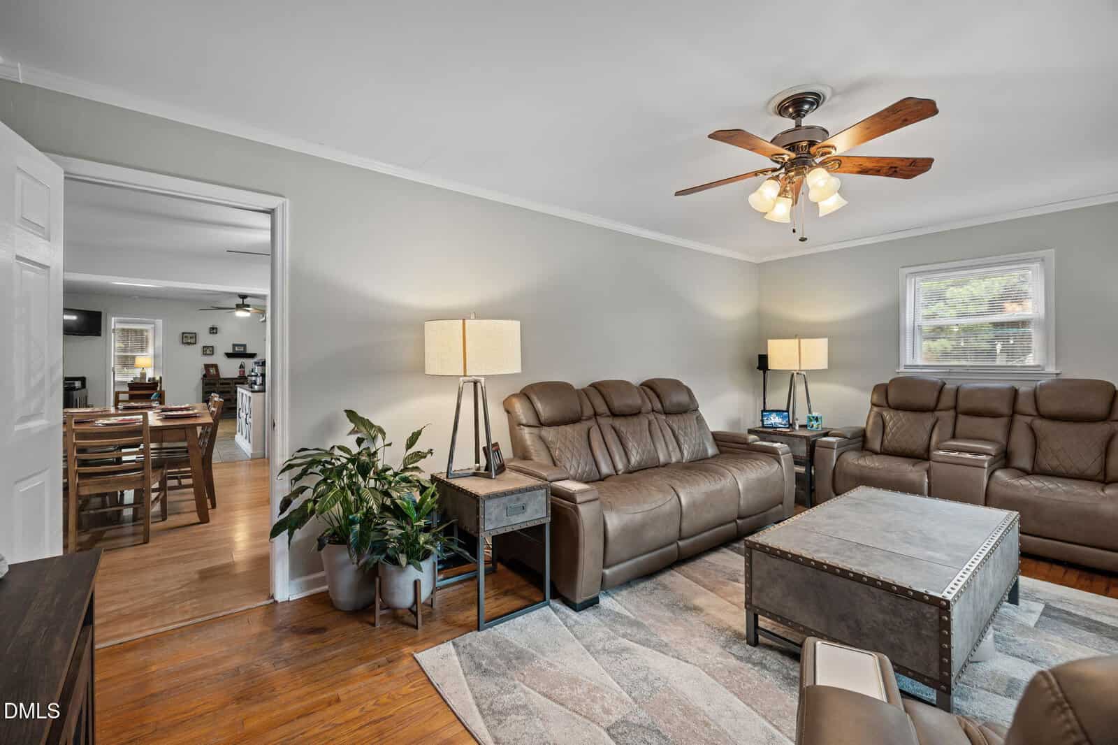 A living room with brown leather sofas, a wooden coffee table, floor and table lamps, potted plants, and a ceiling fan with lights. The room connects to a dining area with a table and chairs visible through an open doorway.