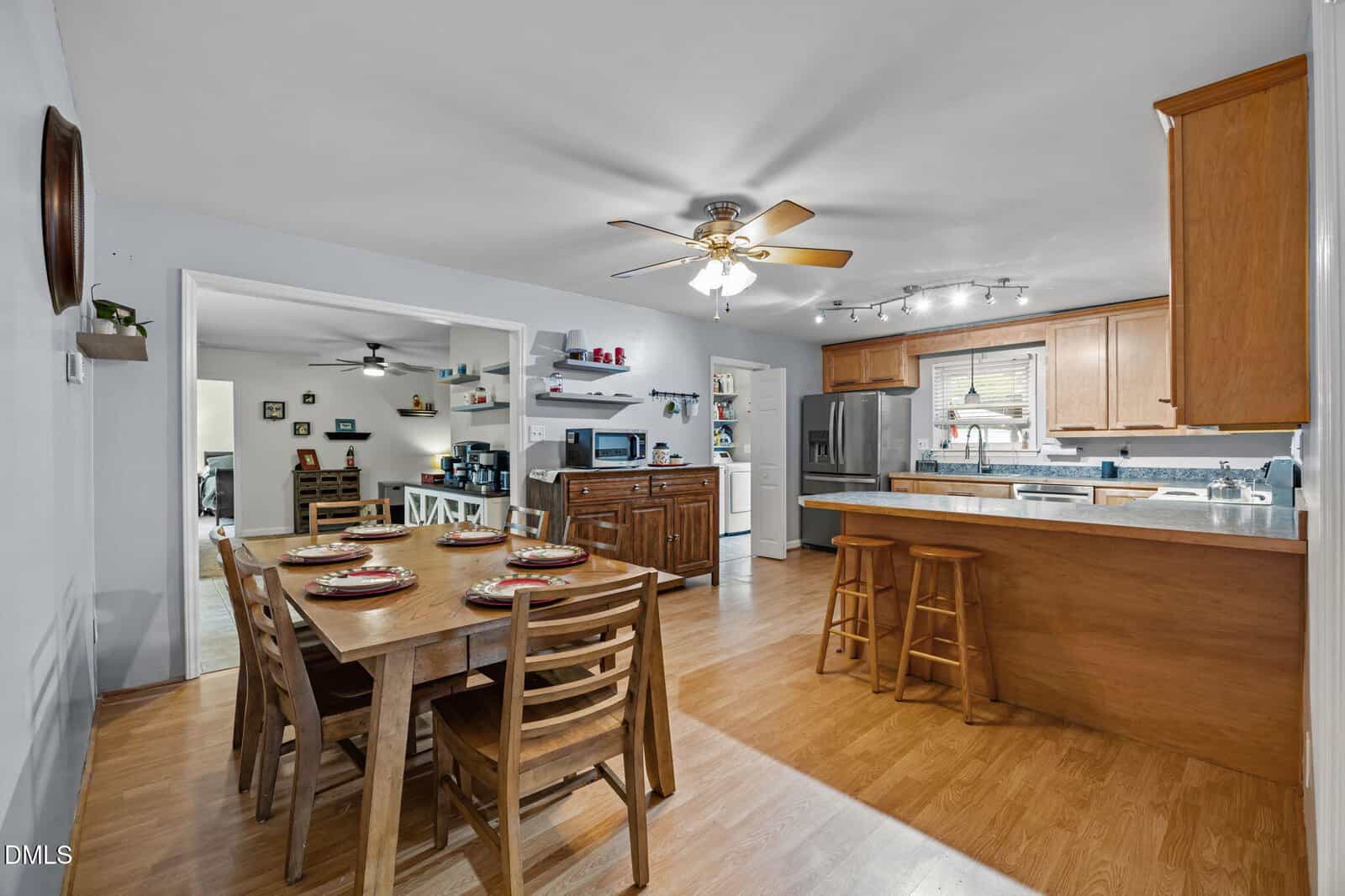 Open kitchen and dining area with wooden floors, a ceiling fan, wooden cabinets, a stainless steel refrigerator, countertop with stools, and a dining table set for four. The space is bright and organized.